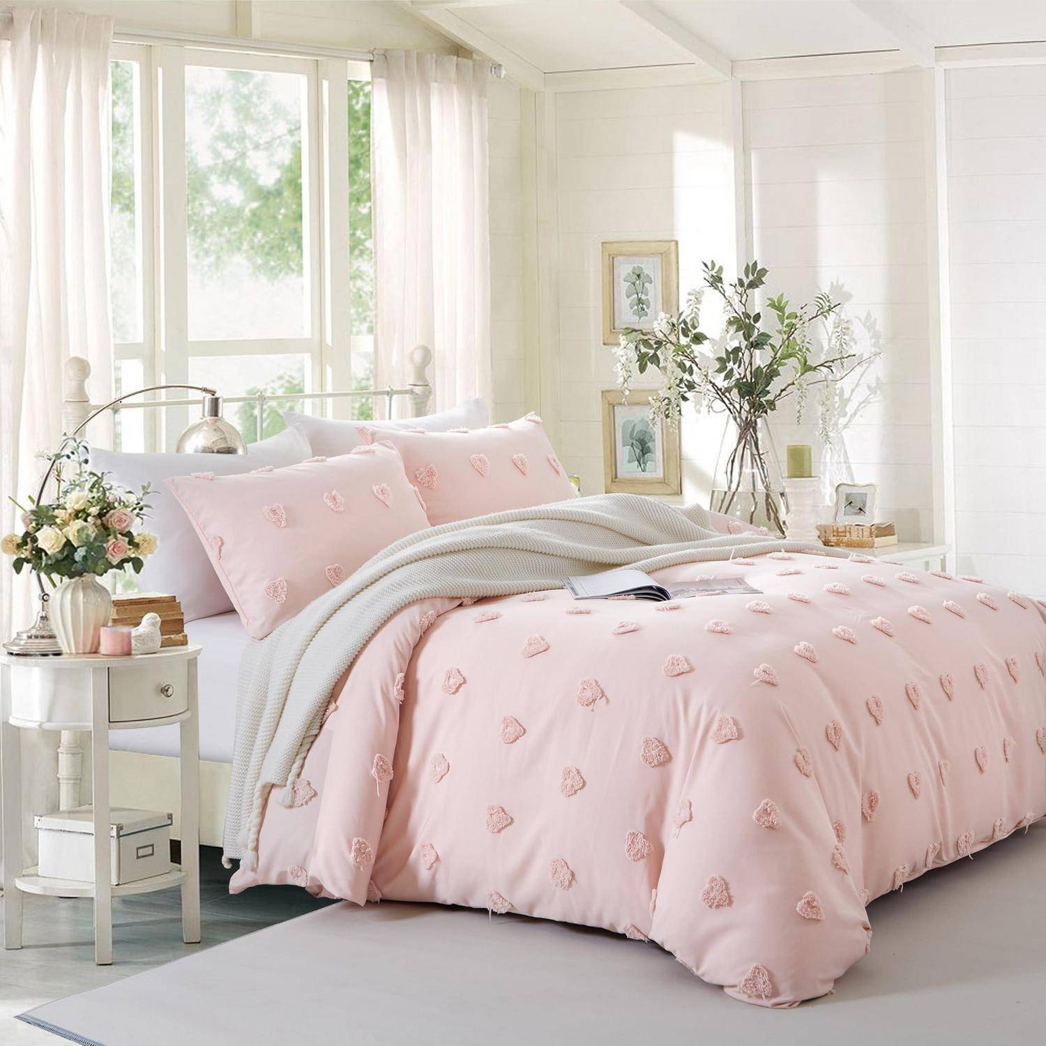 Helles, luftiges Schlafzimmer mit einem ordentlich gemachten Bett in rosa Herzmuster-Bettwäsche, weißen Decken, Nachttisch, Blumen und sonnendurchfluteten Fenstern.