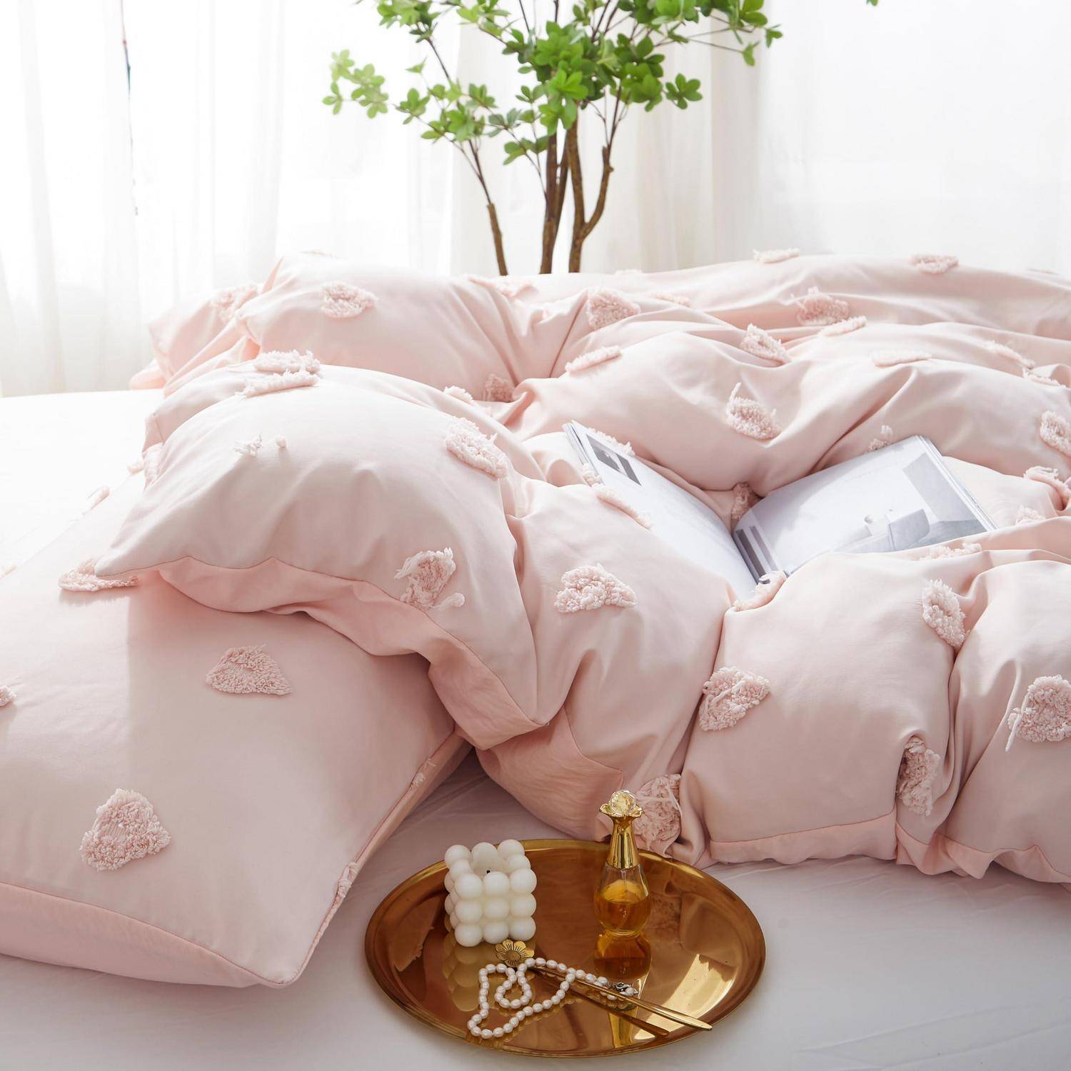 Rosa herzgemusterte Bettwäsche auf einem Bett mit einem goldenen Tablett, auf dem Parfüm, Perlen und eine Kerze stehen, neben einem Fenster mit Pflanzen.