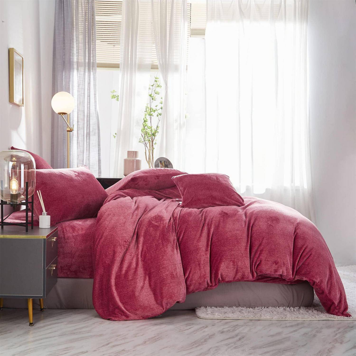 Rosa Bettwäsche und Kissen auf einem Bett neben einem Nachttisch und einer Lampe in einem hellen, modernen Schlafzimmer mit durchsichtigen Vorhängen.