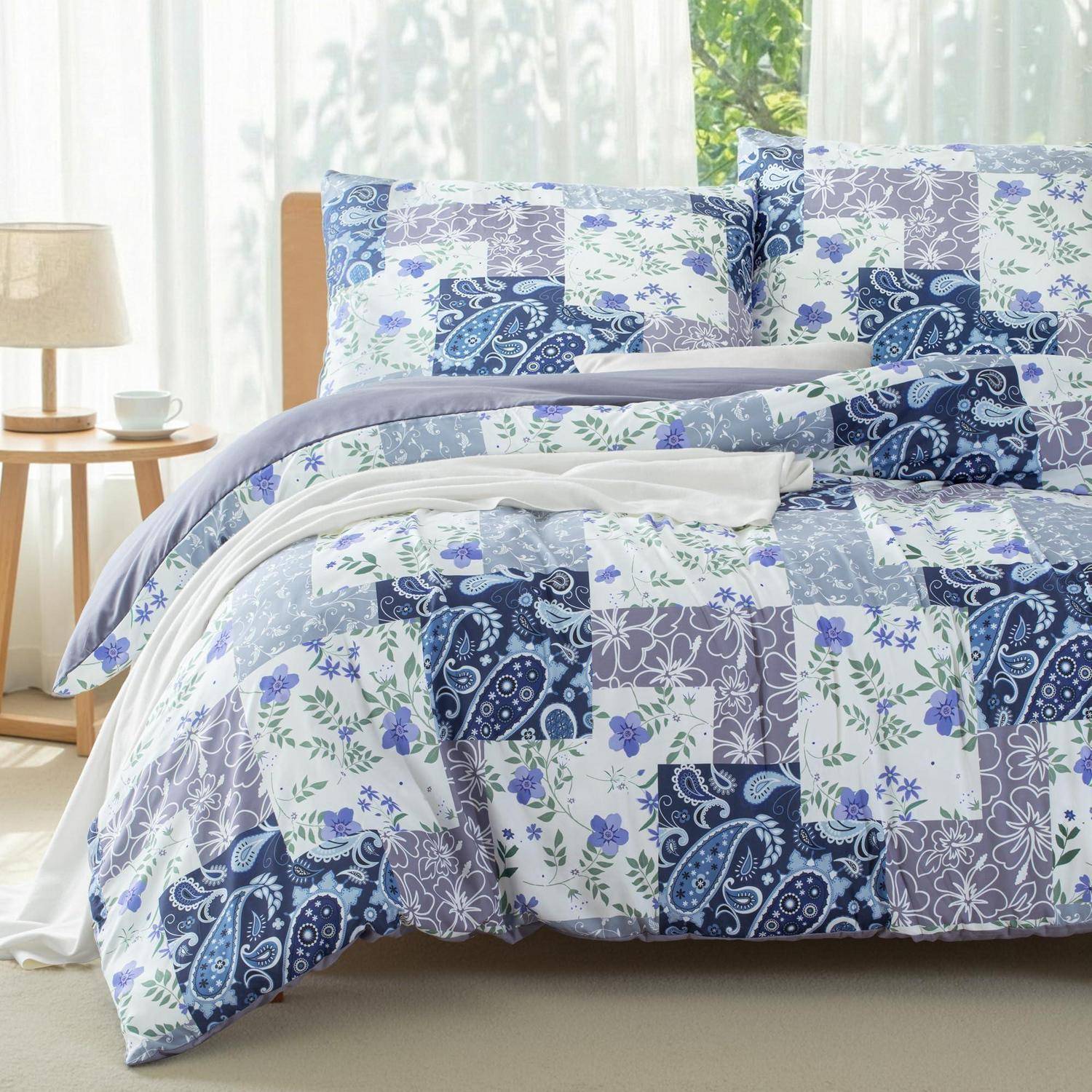 Bett mit blauem und lavendelviolettem Patchwork-Quilt und passenden Kissen in einem hellen Schlafzimmer neben einem Nachttisch und einer Lampe.