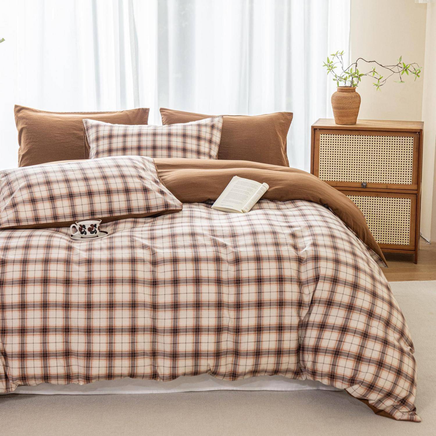 Ordentlich gemachtes Bett mit brauner Karosteppdecke, Kissen, einem aufgeschlagenen Buch und einer Tasse, daneben ein Holzschrank mit einer Vase in einem hellen Schlafzimmer.