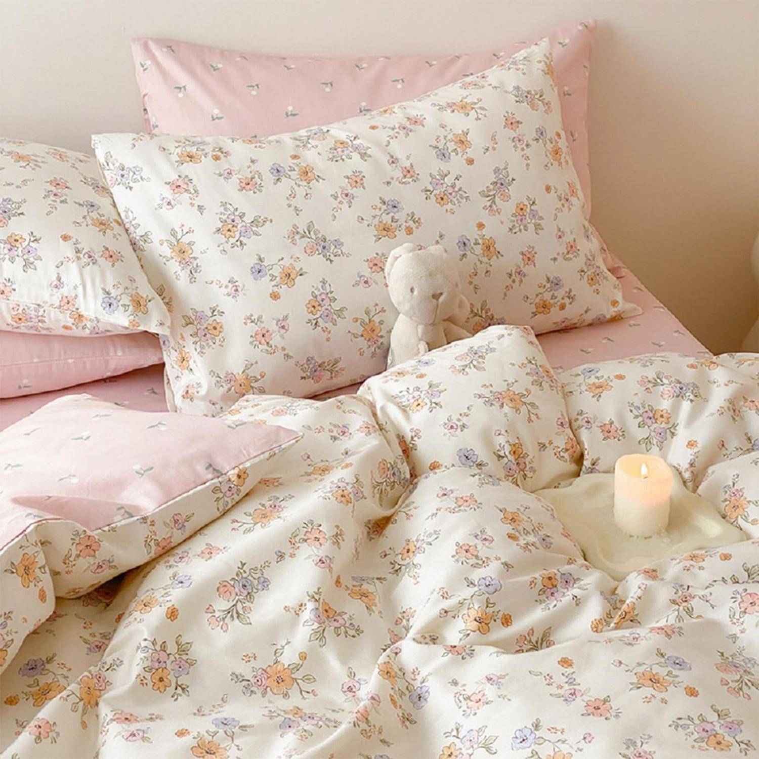 Ordentlich gemachtes Bett mit rosa und blumiger Bettwäsche, mehreren Kissen, einem kleinen Teddy und einer brennenden Kerze, die eine gemütliche Schlafzimmeratmosphäre schafft.