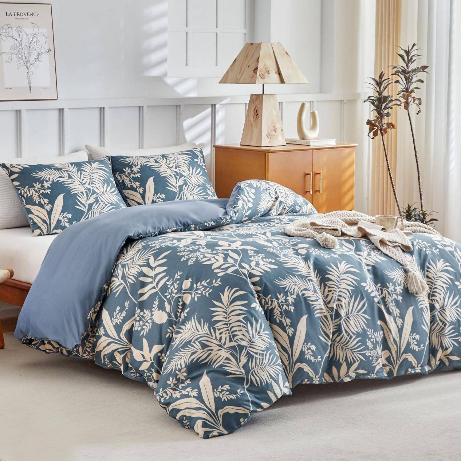 Schlafzimmer mit einem Bett, das in blauer botanischer Bettwäsche und passenden Kissen gekleidet ist, neben einem hölzernen Nachttisch mit Lampe und Dekoration in einem hellen, modernen Innenraum.
