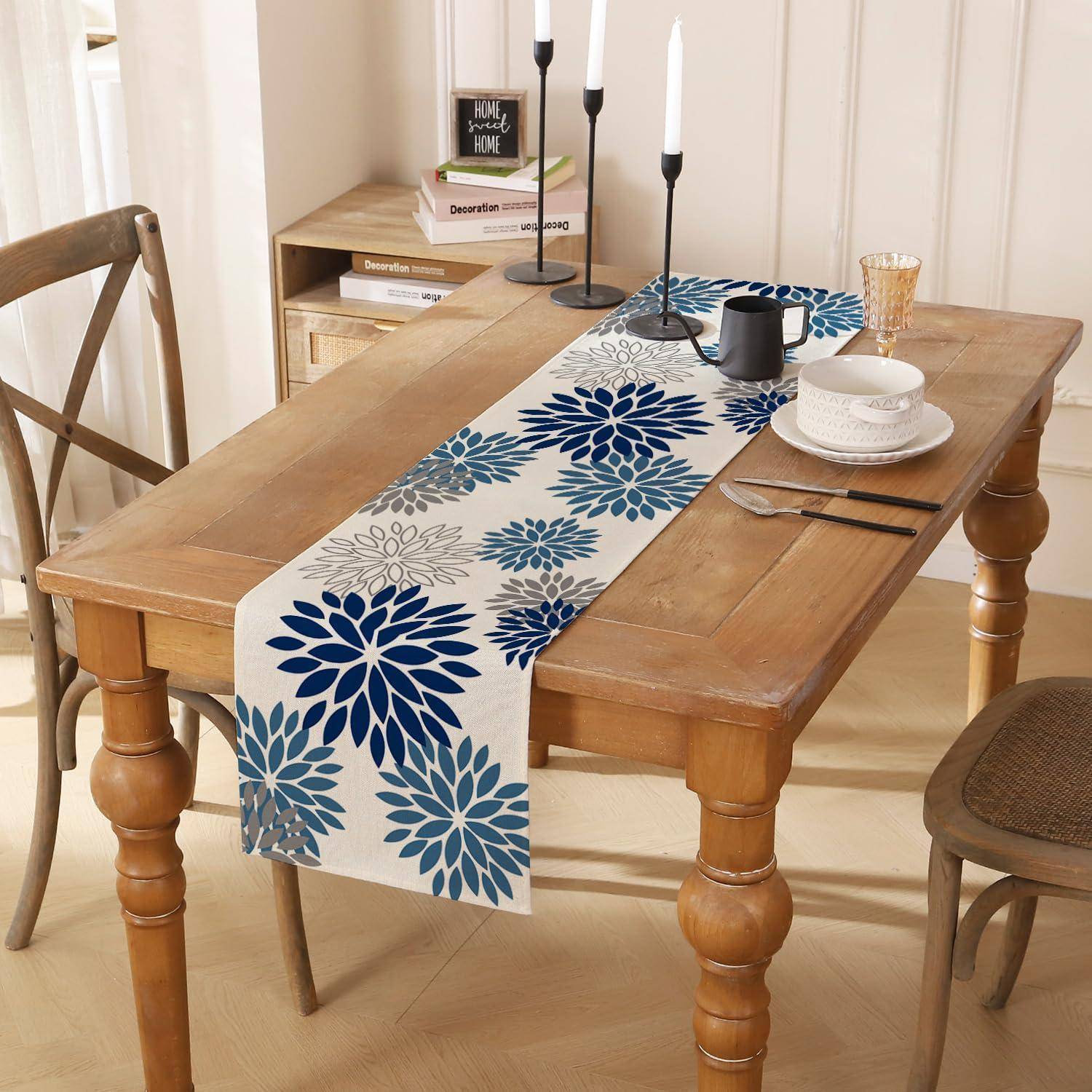 Holztisch zum Essen gedeckt, mit blauem Blumentischläufer, Tellern, Tassen, Besteck, Stühlen und Kerzenhaltern in einem hellen Raum.