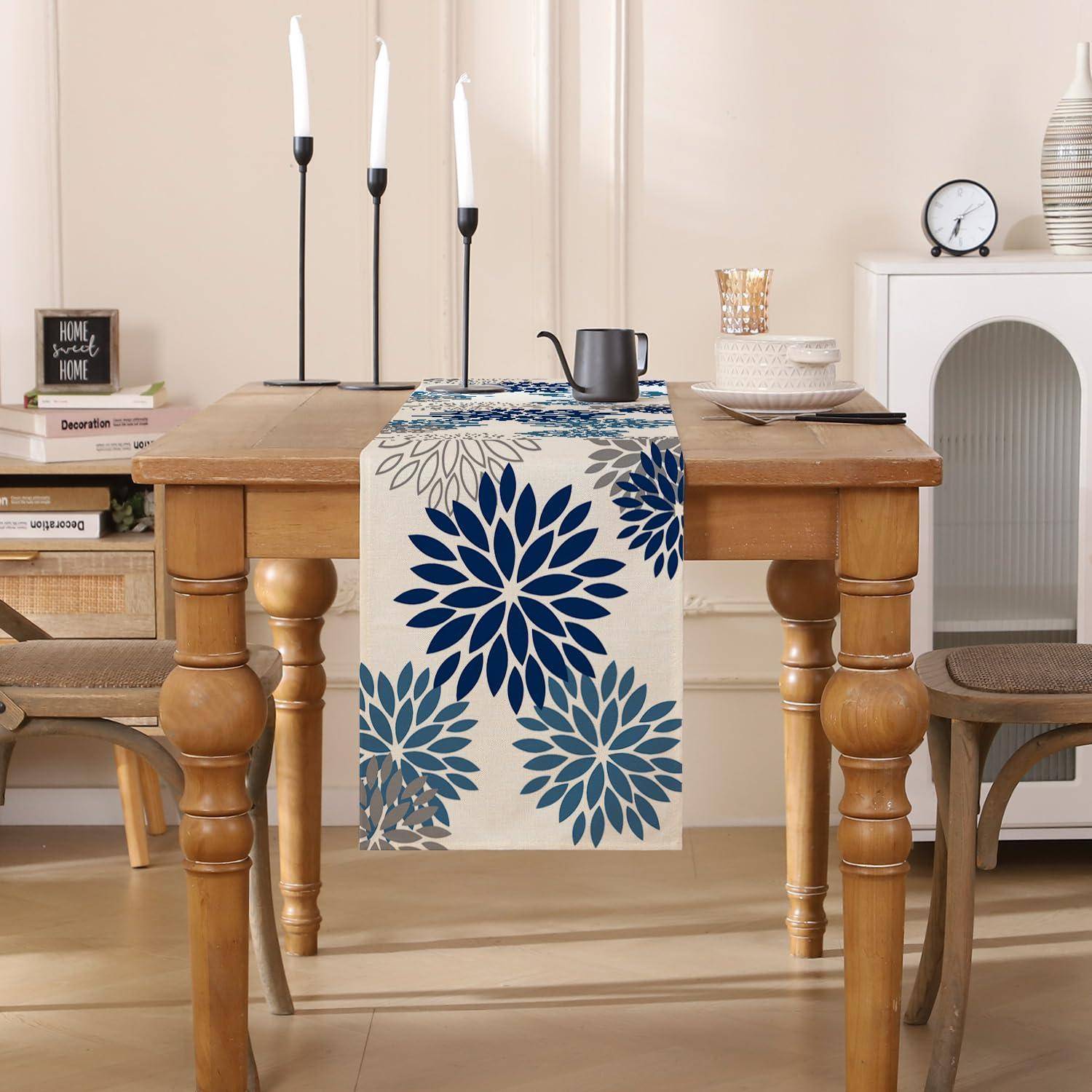 Holztisch mit blauem Blumenlöufer, schwarzen Kerzen, Geschirr und Dekoration in einem beigen Raum mit Stühlen und einem Regal.
