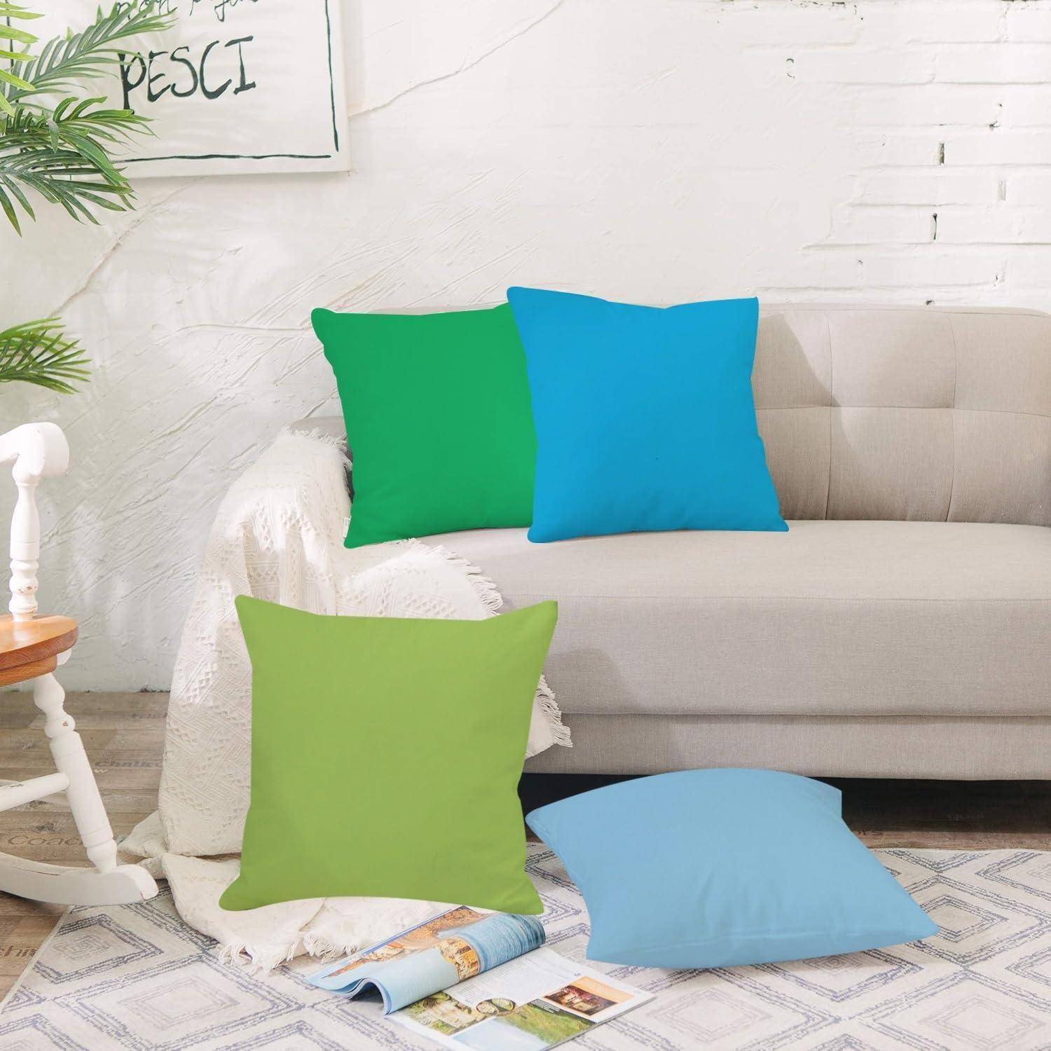 Vier farbenfrohe Dekokissen auf einem beigen Sofa und dem Boden in einem hellen Wohnzimmer, mit einer weißen Decke und einem Teil eines Stuhls sichtbar.