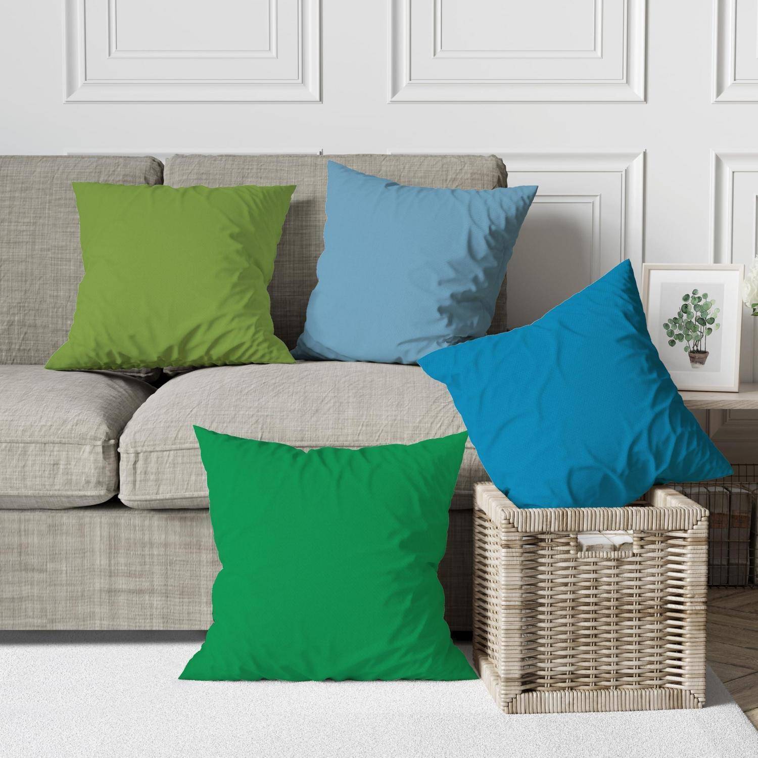 Vier einfarbige Dekokissen in Grün und Blau auf einem beigen Sofa und einem Wicker-Hocker in einem hellen Wohnzimmer angeordnet.