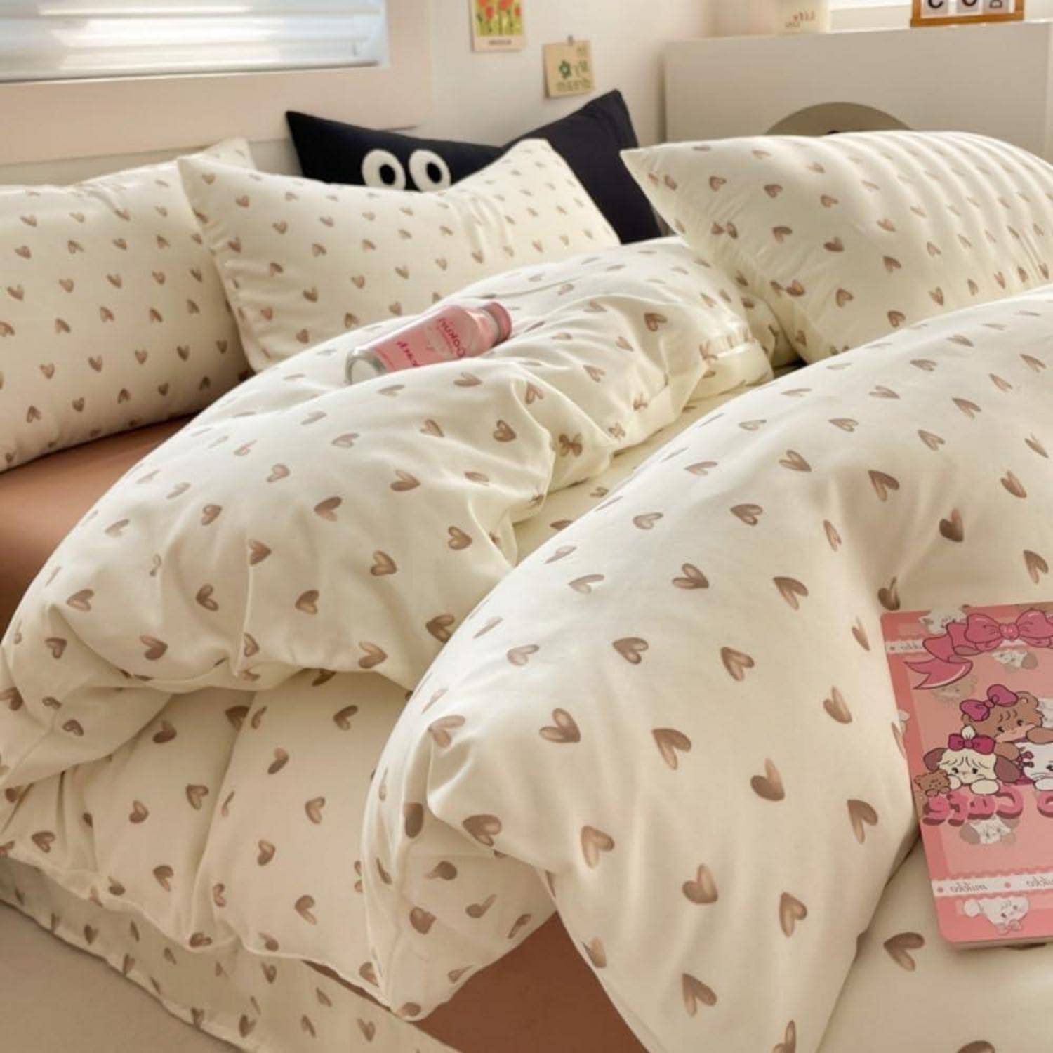 Ungemachtes Bett mit cremefarbenem herzgemustertem Bettzeug und Kissen, eine rosa Flasche auf der Steppdecke und ein kleines rosa Cartoon-Notizbuch am Fußende des Bettes.