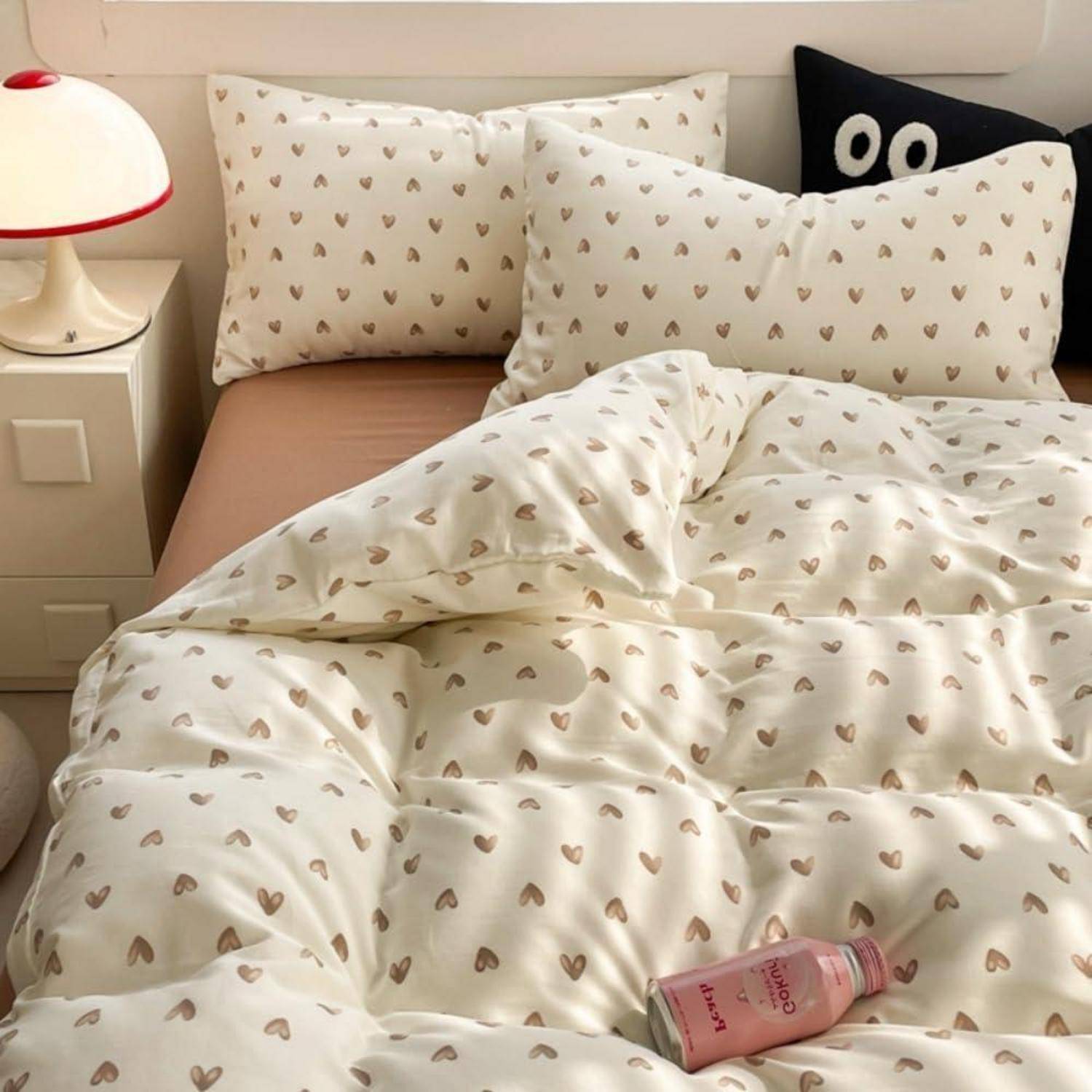 Ein ordentlich gemachtes Bett mit cremefarbenem Bettwäsche- und Kissenmuster mit Herzen, dazu eine rosa Flasche auf der Bettdecke neben einer Nachttischlampe.