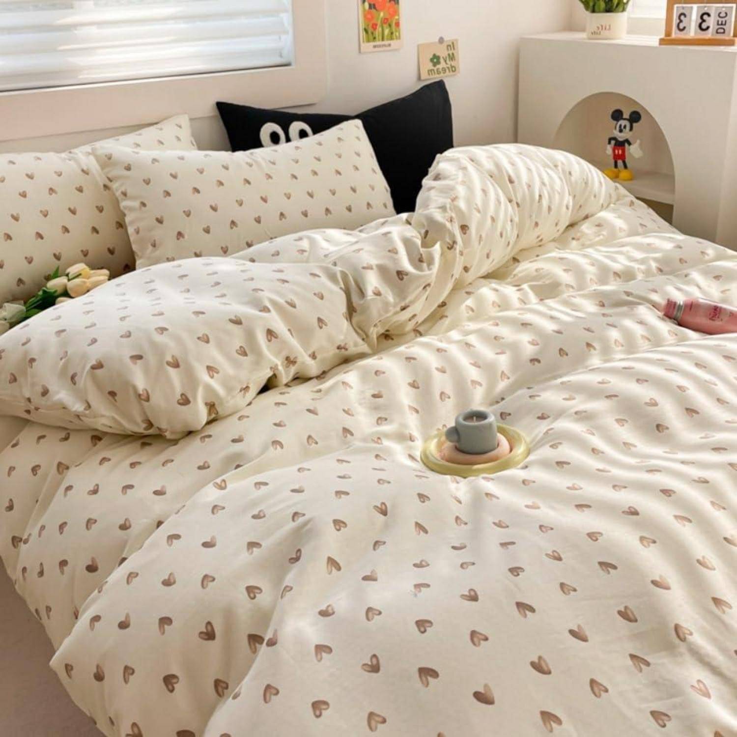 Ungemachtes, unordentliches Bett mit herzförmigem Bettwäsche- und Kissenmuster in einem hellen Schlafzimmer, mit kleinen dekorativen Regalgegenständen und Fensterjalousien.