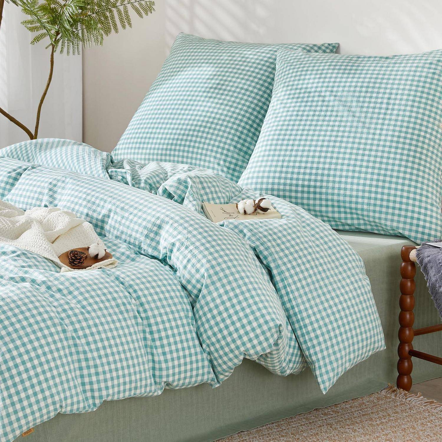 Ein Bett mit mintgrüner Gingham-Bettwäsche, bestehend aus Kissen und einer Steppdecke, gestaltet in einem hellen Schlafzimmer mit einer Pflanze in der Nähe.