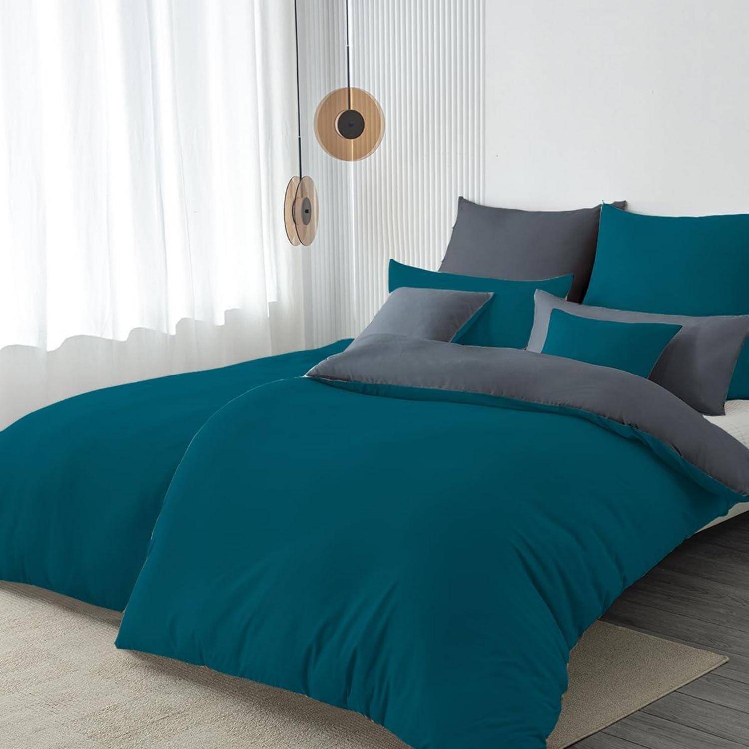Ein ordentlich gemachtes Bett mit blaugrüner und grauer Bettwäsche in einem hellen, modernen Schlafzimmer neben einem Fenster mit durchscheinenden Vorhängen.