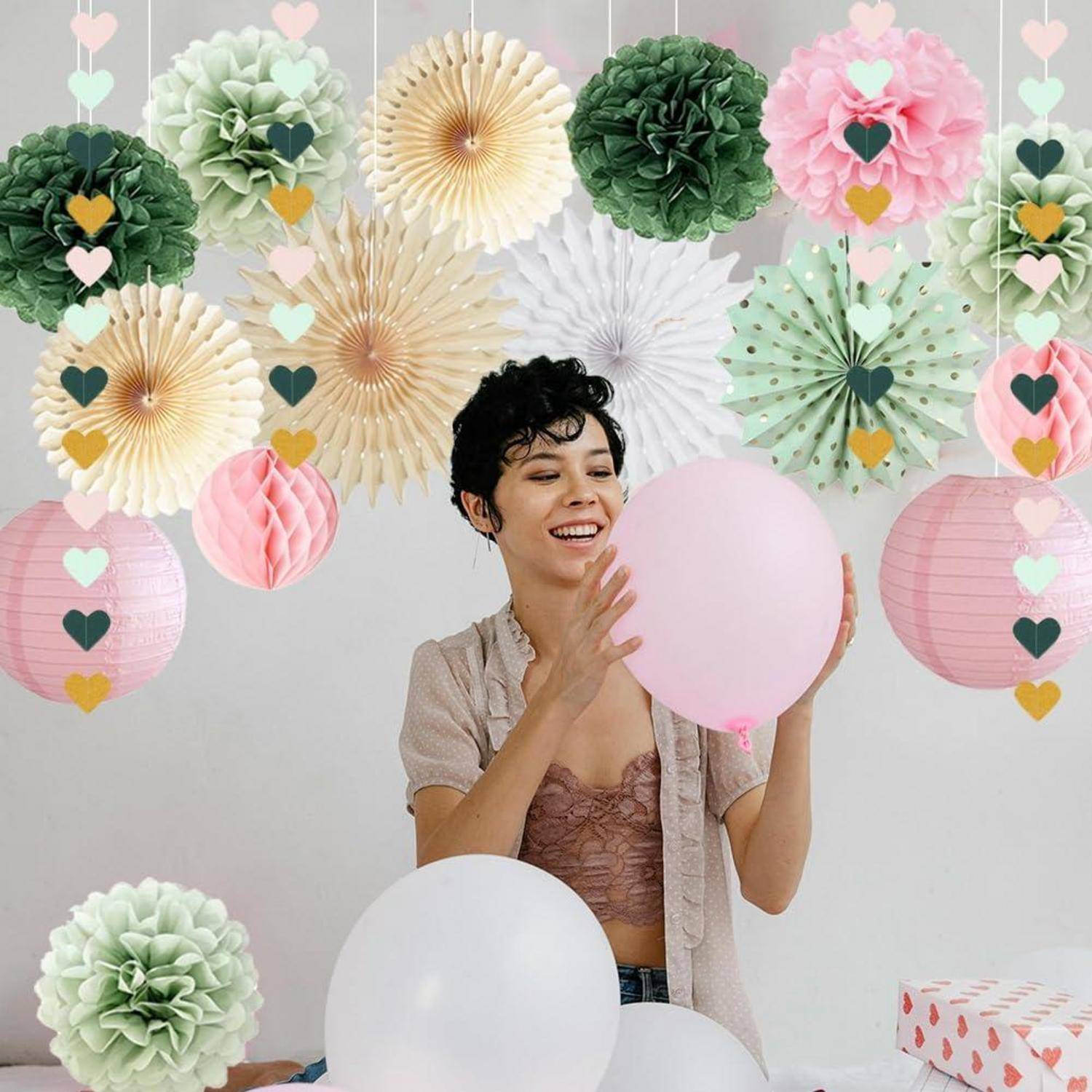 Lächelnde Frau vor einer Pastellparty-Kulisse hält einen rosa Ballon und ist umgeben von hängenden Papierblumen, Herzgirlanden und Ballons.