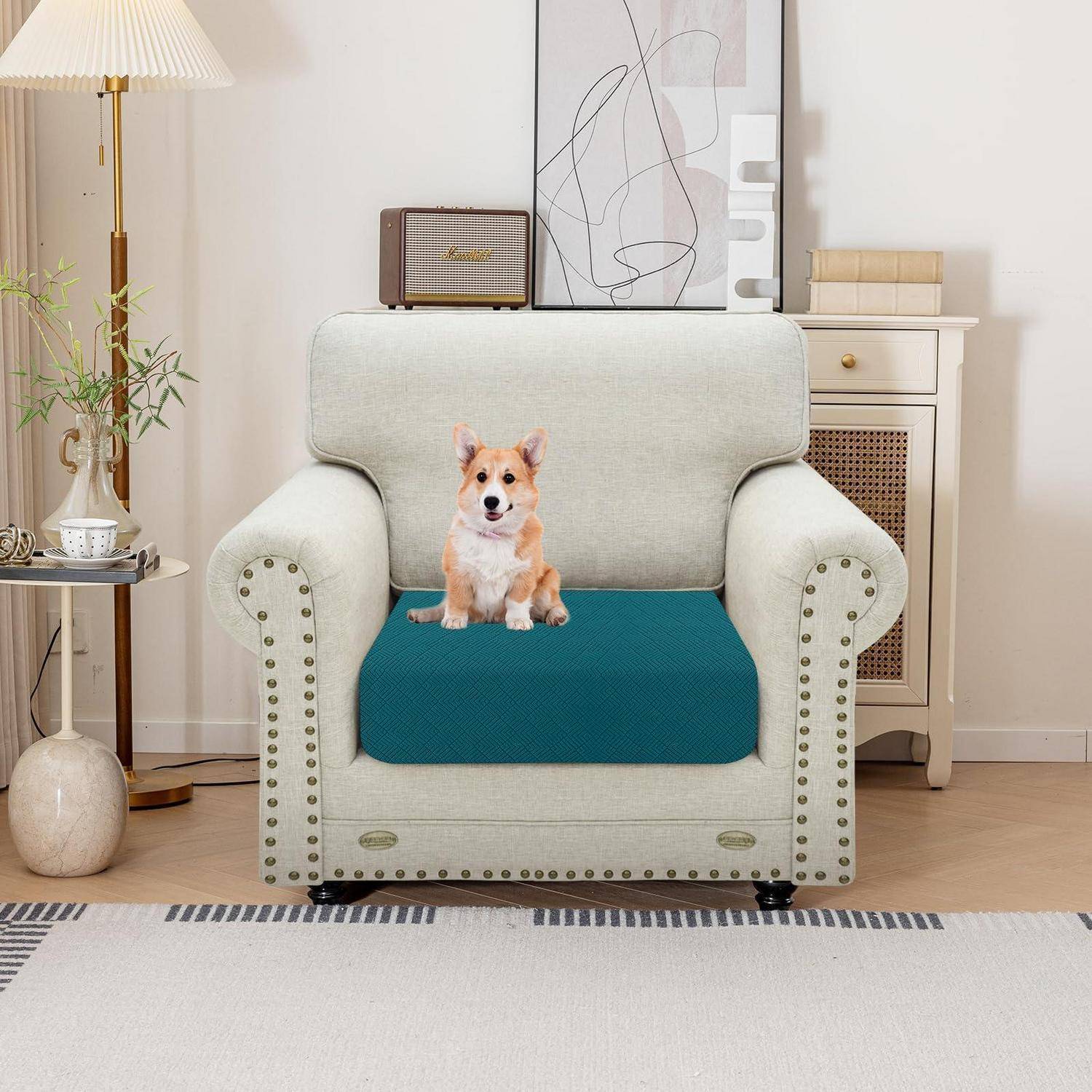 Ein kleiner Corgi sitzt auf einem blaugrünen Kissen, das auf einem beigefarbenen Sessel in einem gemütlichen Wohnzimmer platziert ist.