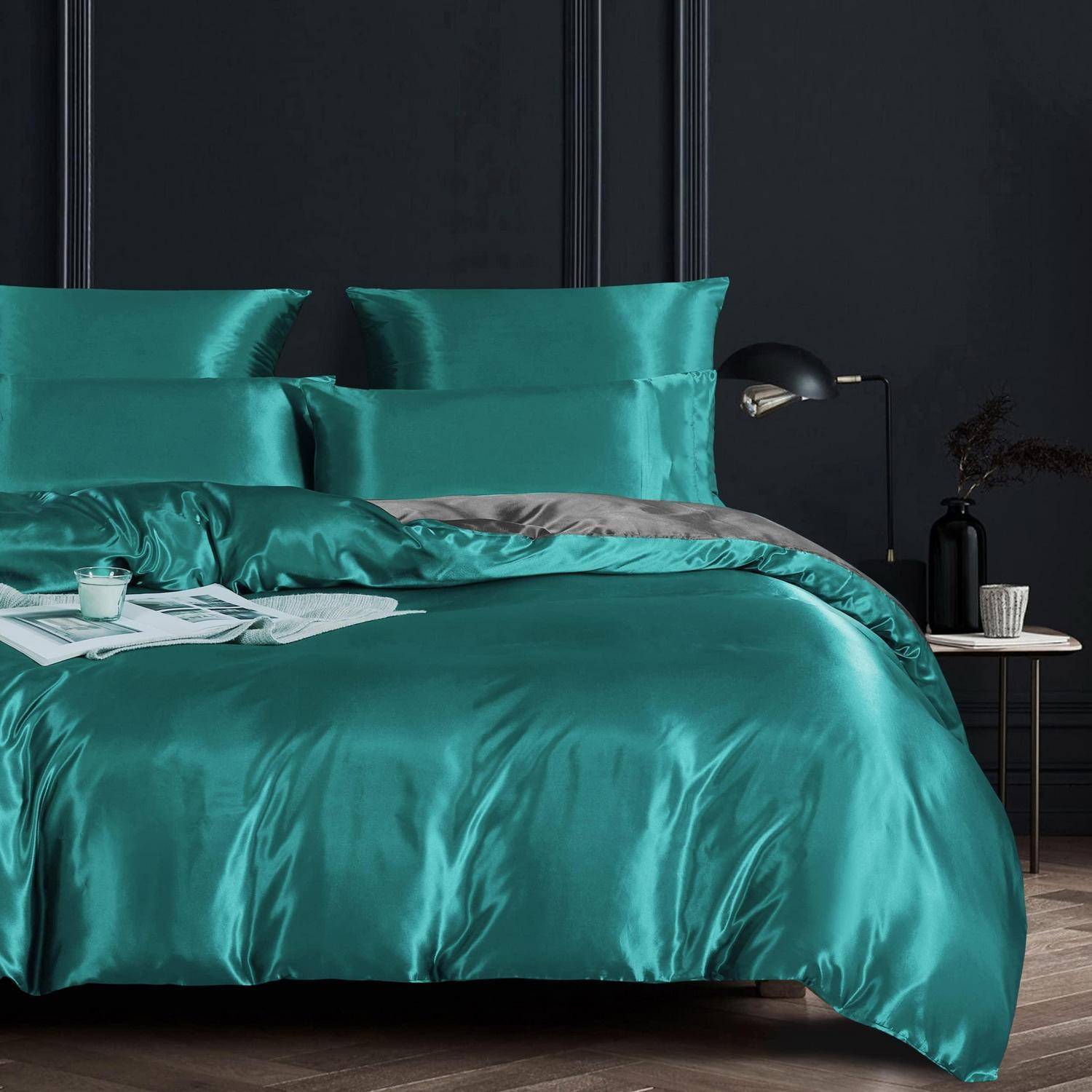Tealfarbene Satinbettwäsche auf einem ordentlich gemachten Bett in einem dunklen, modernen Schlafzimmer, mit Kissen, einer Wurfdecke, Büchern und einer Nachttischlampe.