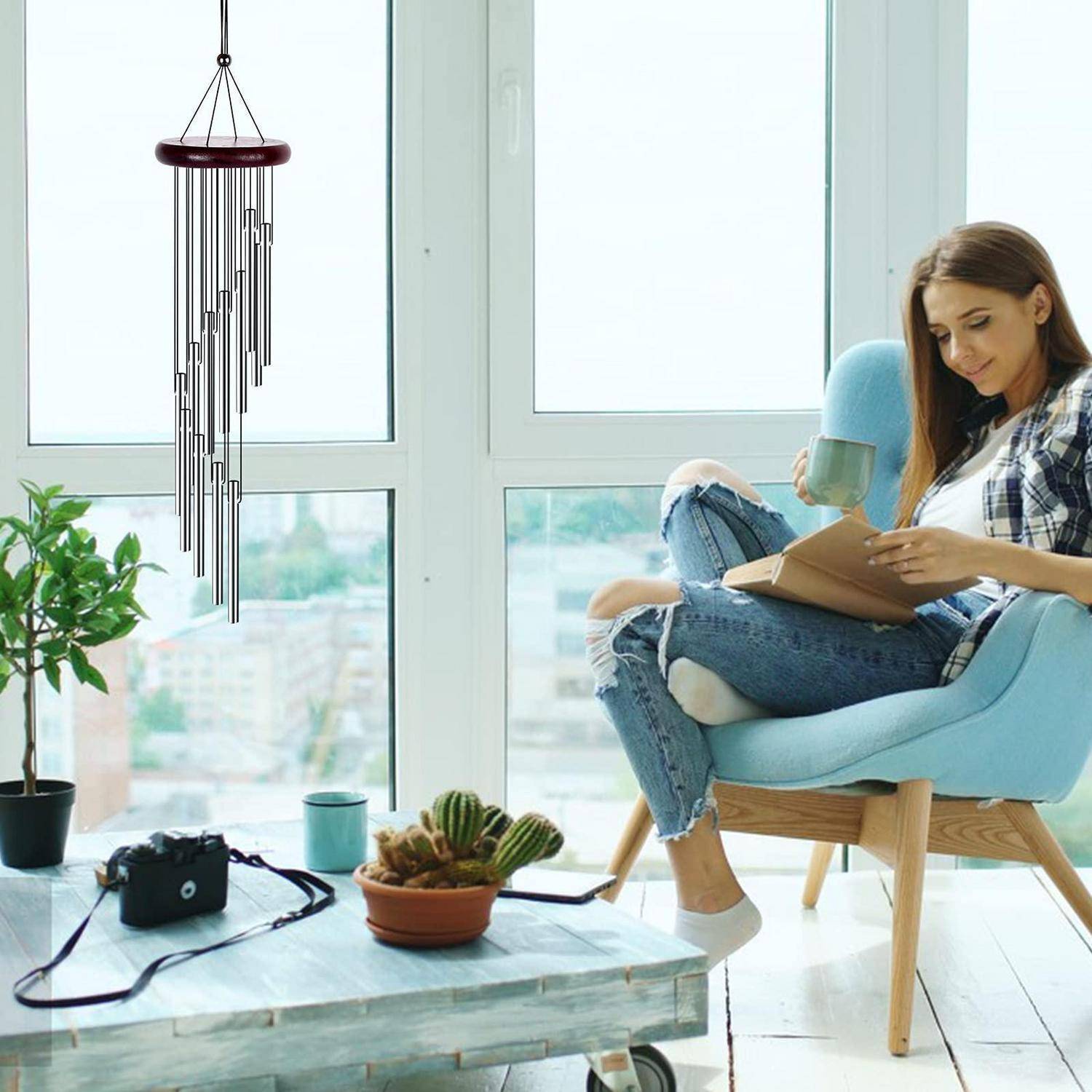 Eine Frau entspannt sich in einem Stuhl neben einem Fenster, liest ein Buch und hält eine Tasse in der Hand; eine Kamera, Pflanzen und ein Windspiel sind in der Nähe.