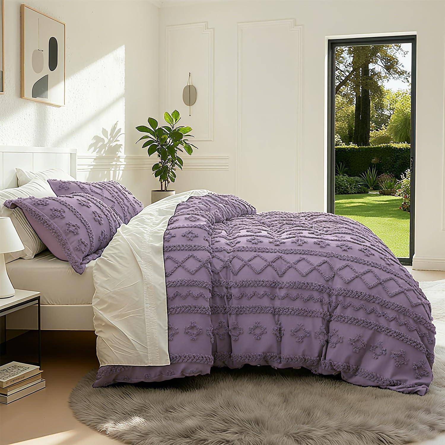 Ein ordentlich gemachtes Bett mit einer strukturierten Lavendeldecke und Kissen in einem hell weißen Schlafzimmer, mit einer Topfpflanze und Gartenblick durch eine offene Türöffnung.