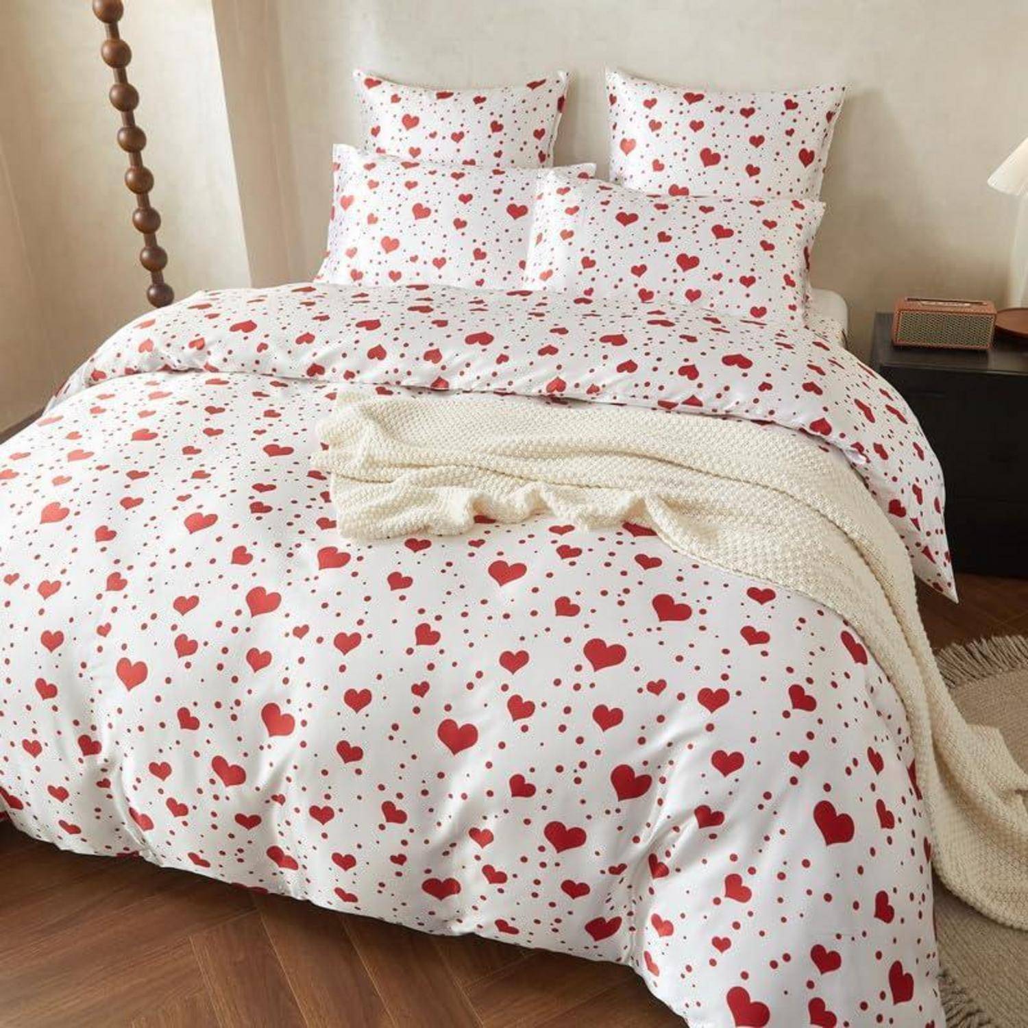 Ein Bett mit weißer Bettwäsche mit roten Herzdrucken, versehen mit Kissen und einer cremefarbenen Strickdecke, in einem gemütlichen Schlafzimmer.
