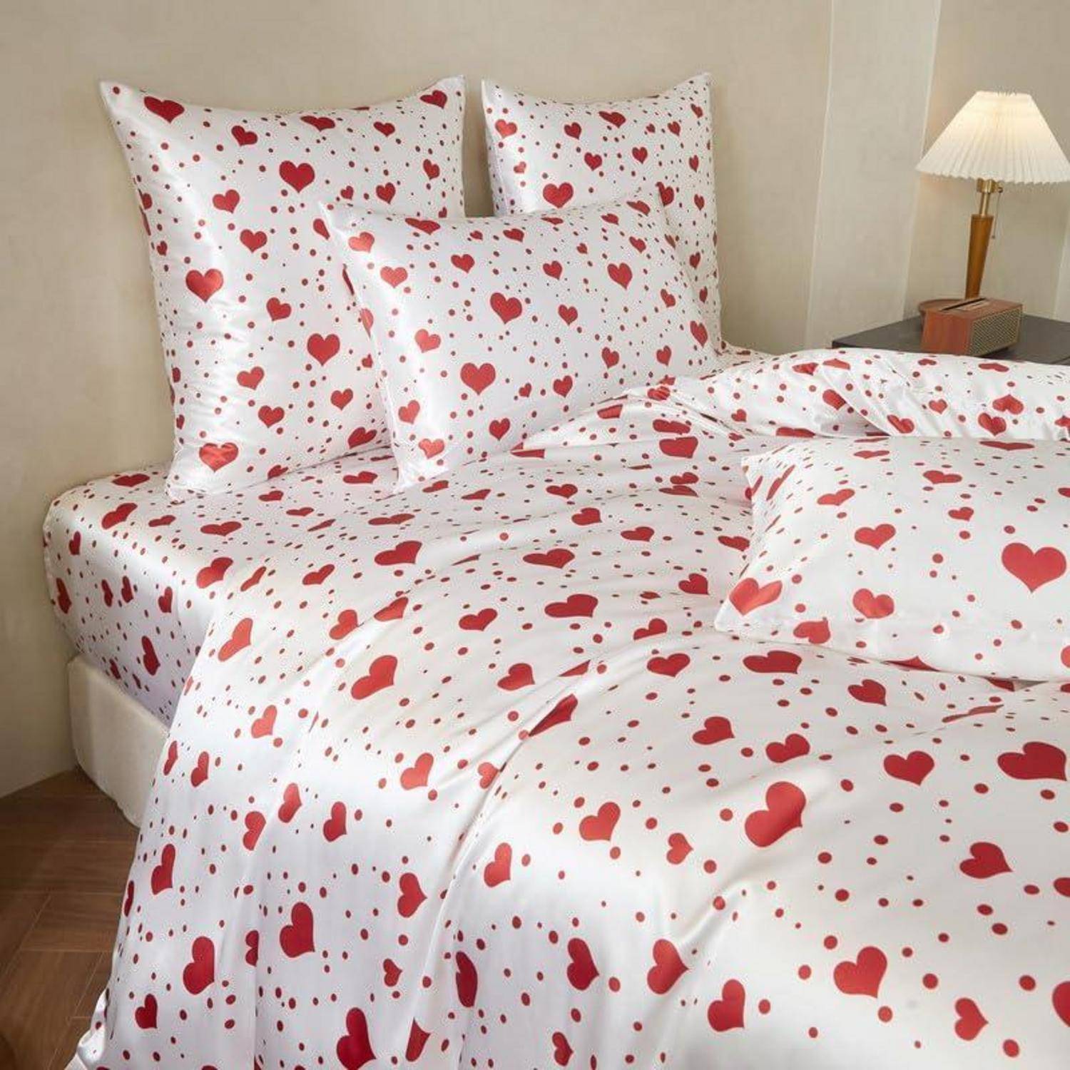Bett mit weißer Bettwäsche, die mit roten Herzen bedeckt ist, einschließlich passender Kissen und einer zusammengefalteten Decke in einem Schlafzimmer.