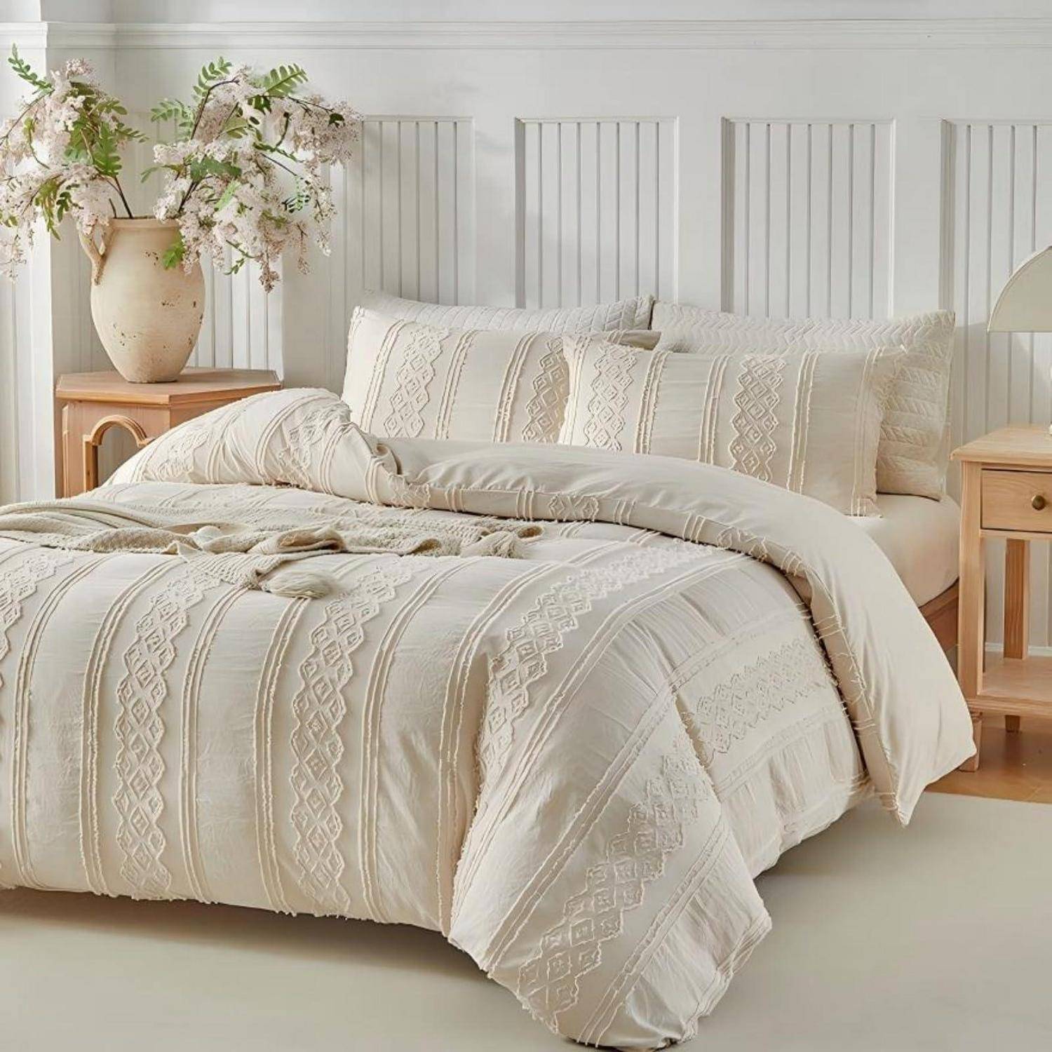 Cremefarbenes Boho-Bett mit strukturierter gemusterter Bettdecke und Kissen, umrahmt von weißem Paneele-Kopfteil und kleinen Holz-Nachttischen mit Blumen.