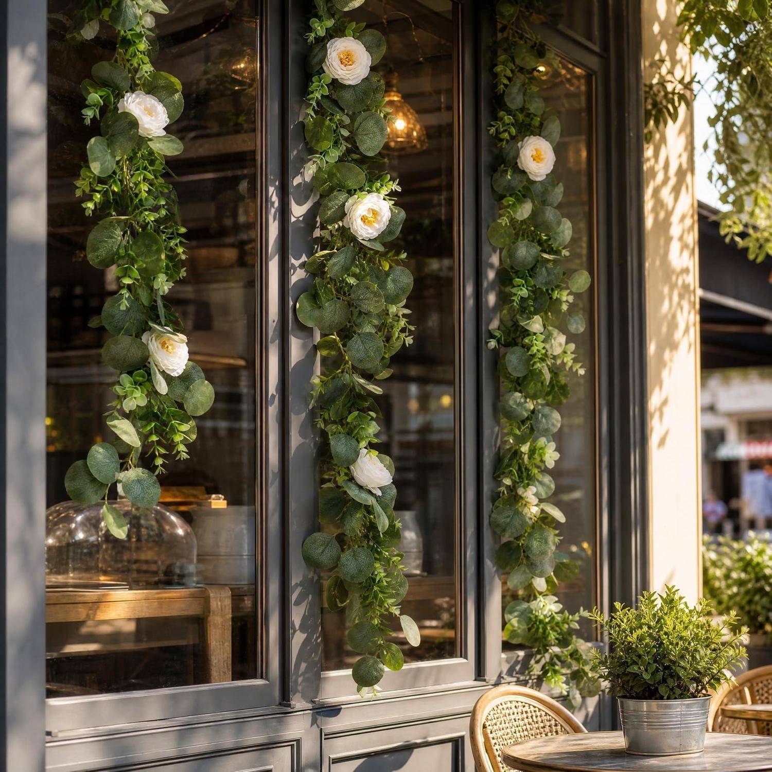 Blumenkränze mit weißen Blüten hängen an einem Café-Fenster neben Korbmöbeln und Topfpflanzen im warmen Tageslicht.