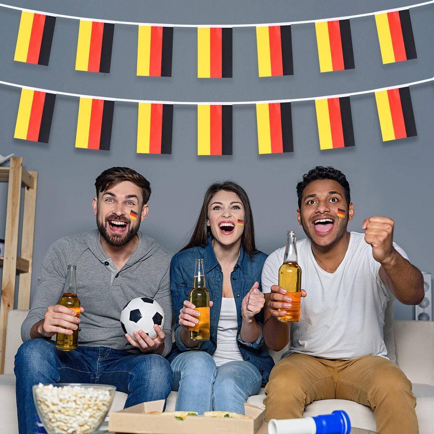 Drei Freunde auf einem Sofa jubeln für Deutschlands Mannschaft, halten Bier und einen Fußball unter deutscher Flaggengirlande.