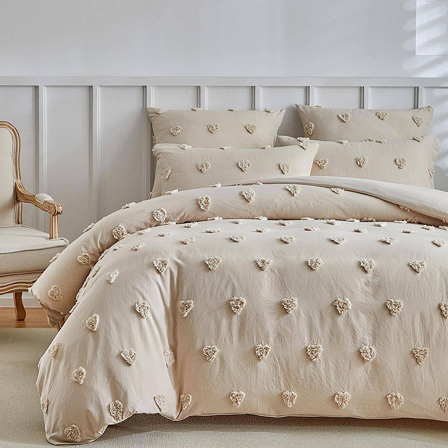 Ein ordentlich gemachtes Bett mit beigefarbener Bettwäsche mit Herzenmuster und Kissen in einem hellen, minimalistischen Schlafzimmer neben einem Holzstuhl.