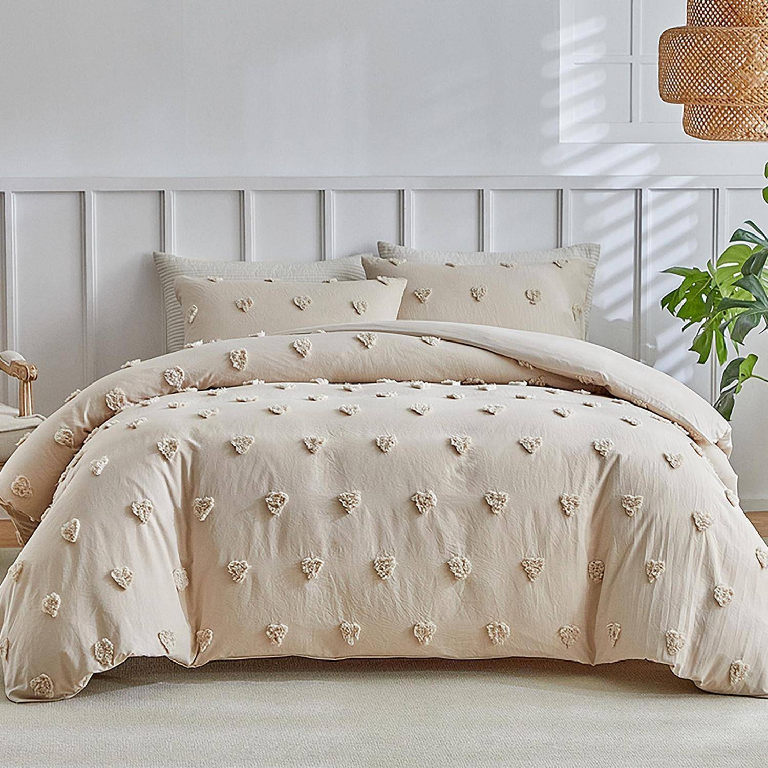 Beiges Bett mit herzgemusterter Bettdecke und Kissen in einem hellen, minimalistischen Schlafzimmer mit weißem Kopfteil und Pflanze.