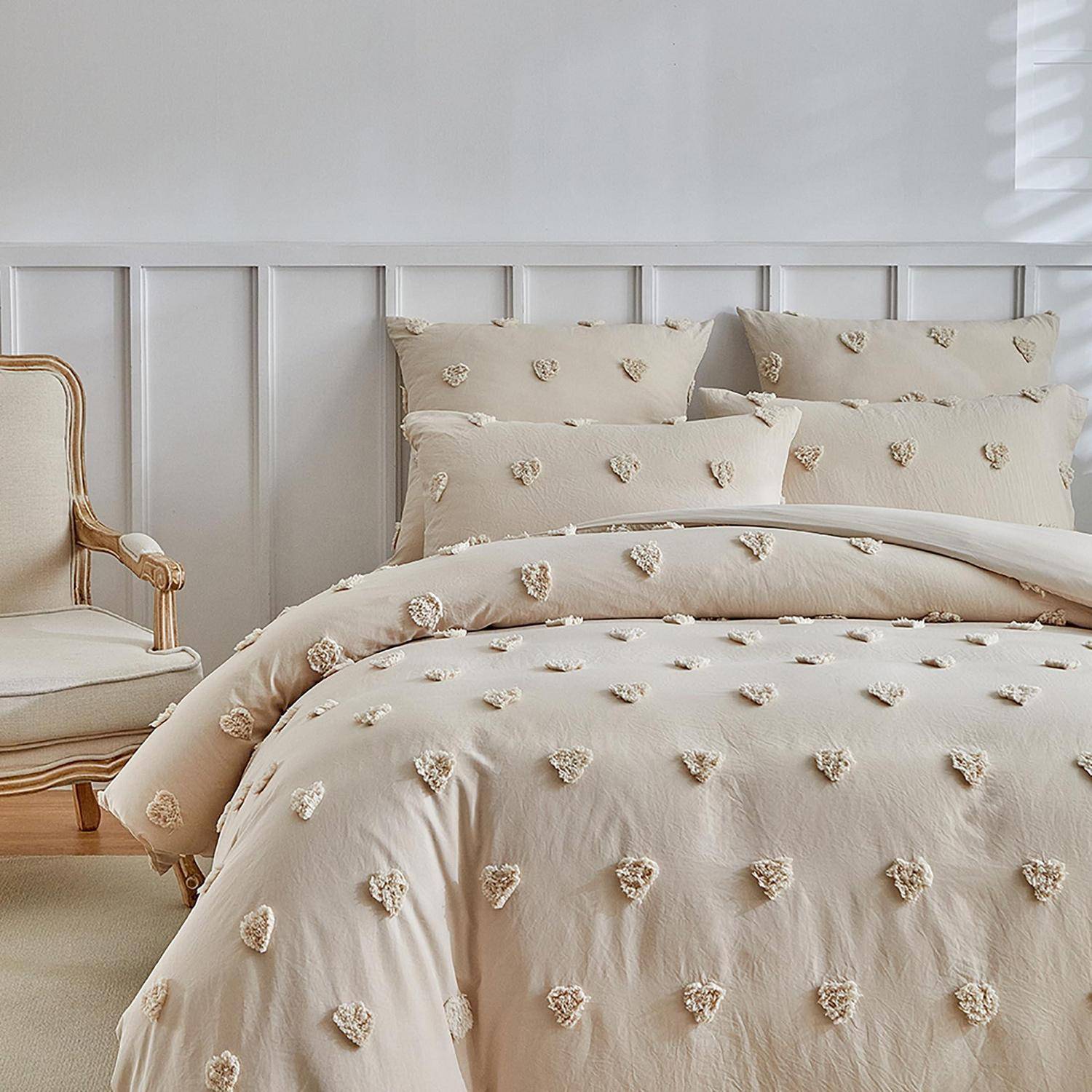 Beiges strukturiertes Bett mit passenden Kissen und Bettdecke in einem hellen Schlafzimmer neben einem gepolsterten Sessel und weißer Täfelwand.