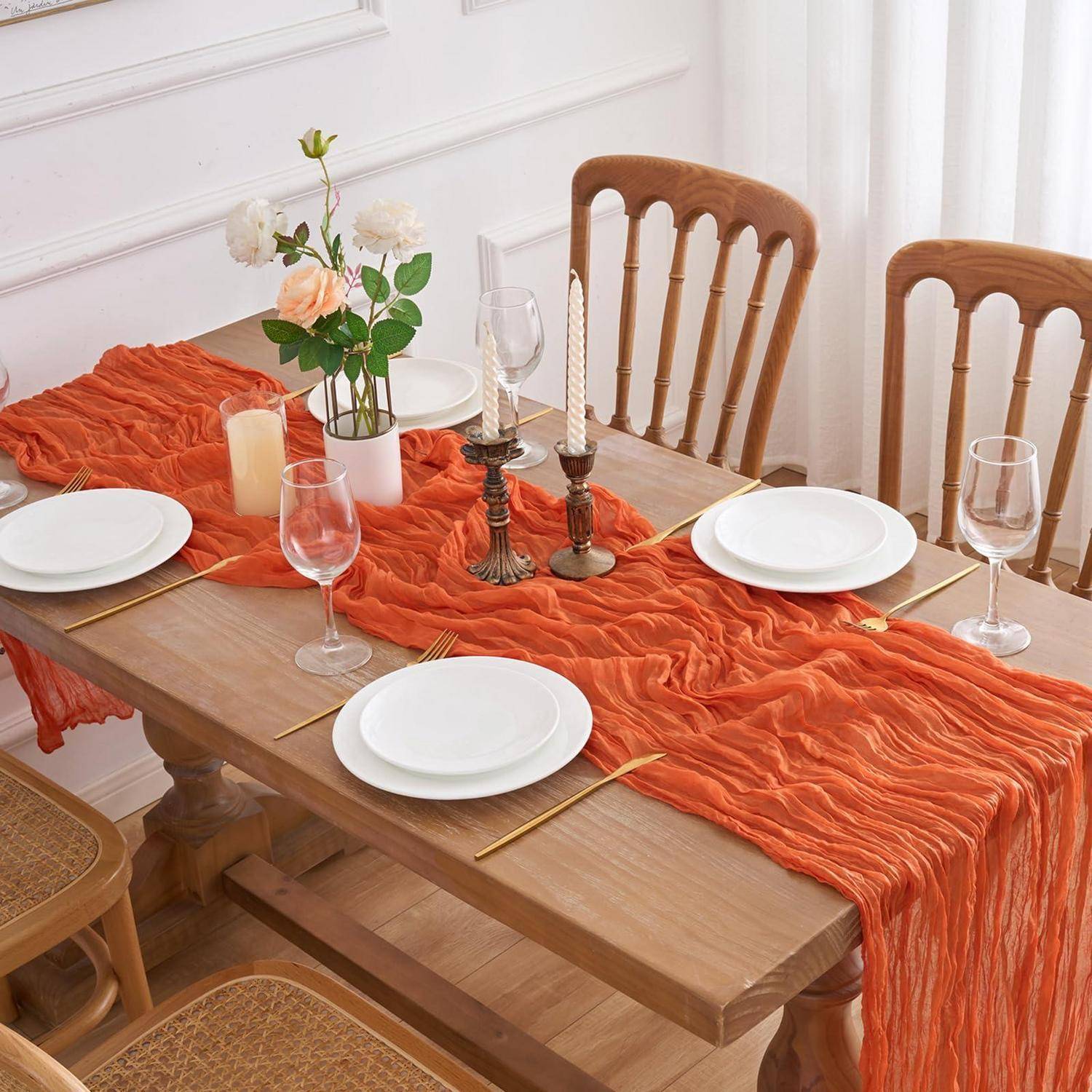 Ein eingedeckter Esstisch mit einem orangefarbenen strukturierten Tischläufer, weißen Tellern, Weingläsern, Kerzen und einer kleinen Vase mit Blumen in einem hellen Raum.
