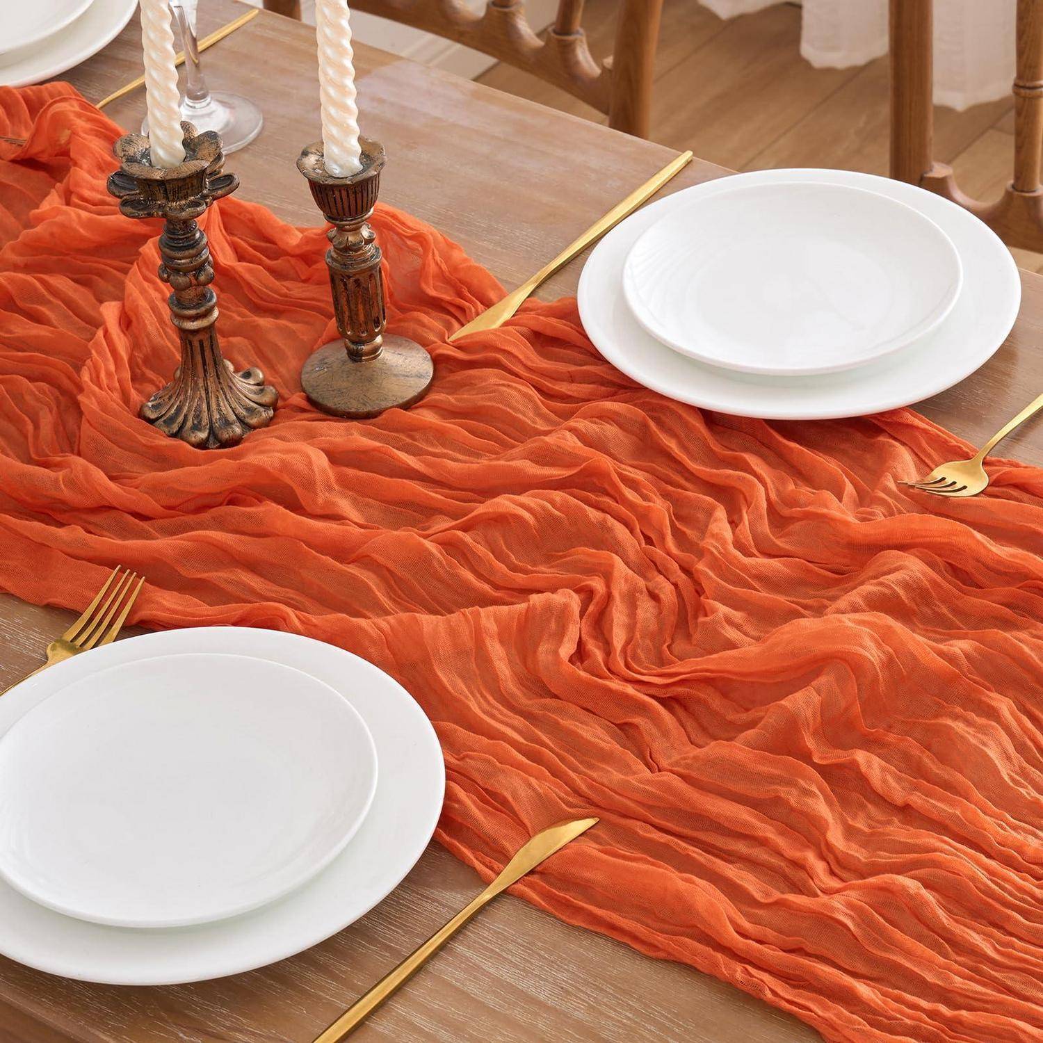 Decken Sie den Esstisch mit weißen Tellern, goldfarbenen Bestecken und zwei Kerzenleuchtern auf einem orangefarbenen Tischläufer mit Knitteroptik.