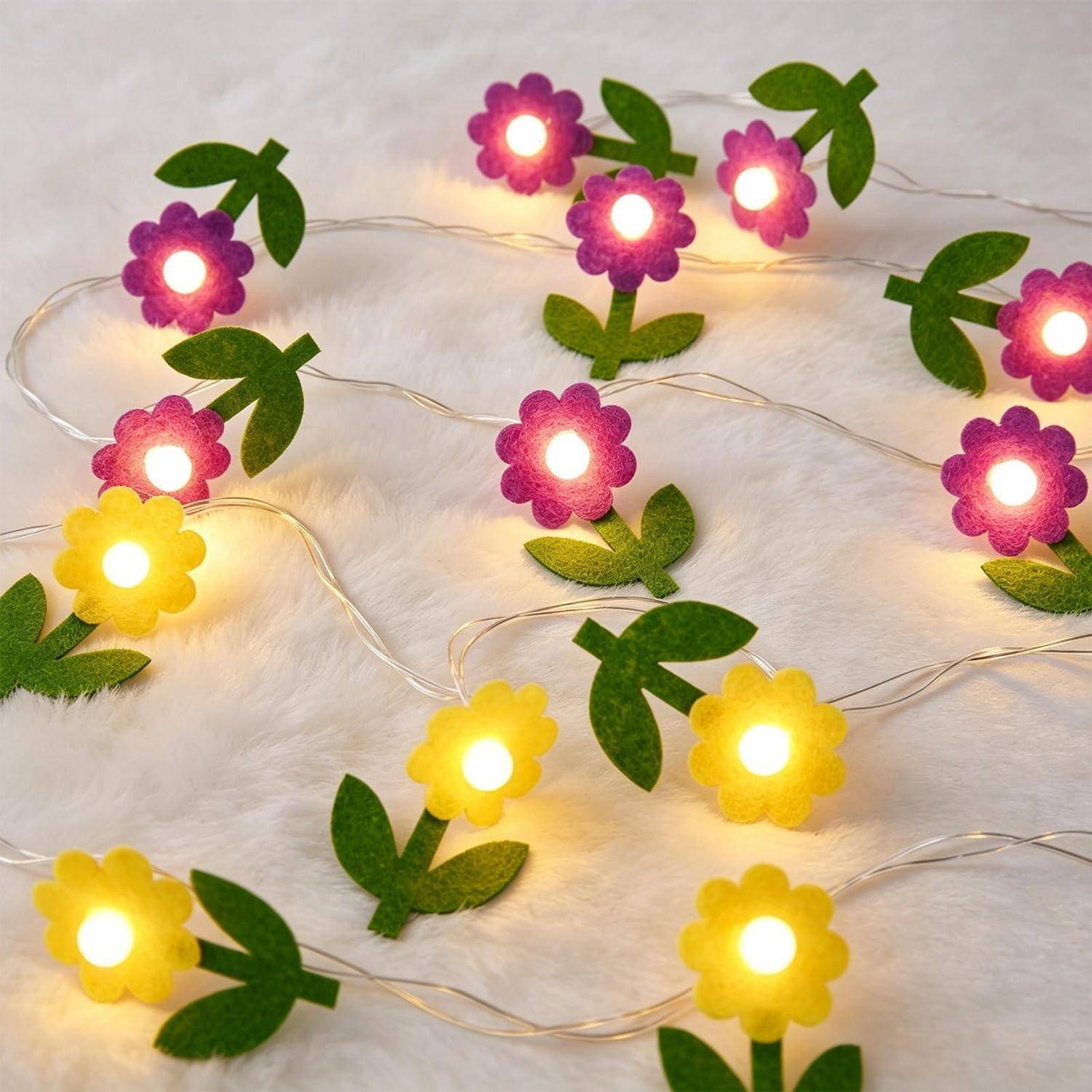 Blütenförmige Lichterketten in Lila und Gelb mit grünen Blättern, leuchtend auf weißem flauschigem Hintergrund.