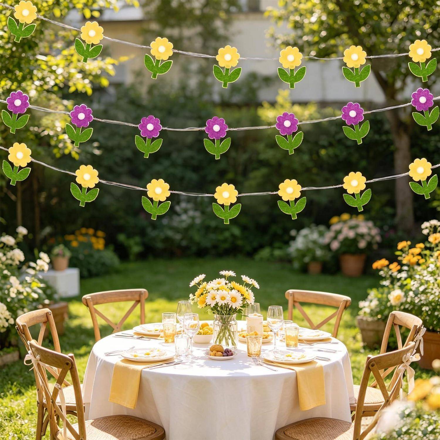 Außengartenparty-Setup mit einem runden Tisch für vier Personen, weißer Tischdecke, gelben Akzenten, Blumen und hängenden Blumengirlanden über dem Kopf.