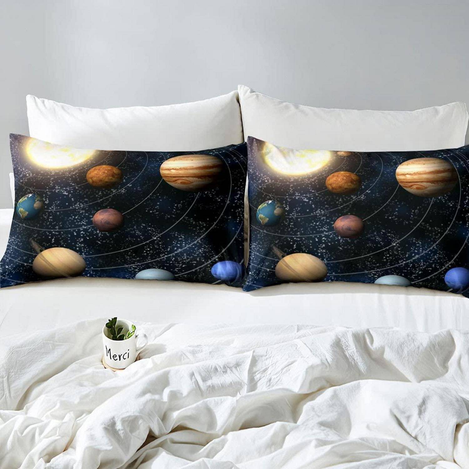 Zwei weltraum-thematische Dekokissen mit Sonnensystem-Druck auf einem weißen Bett mit zerknitterter weißer Bettwäsche und einem kleinen "Merci""-Blumentopf.