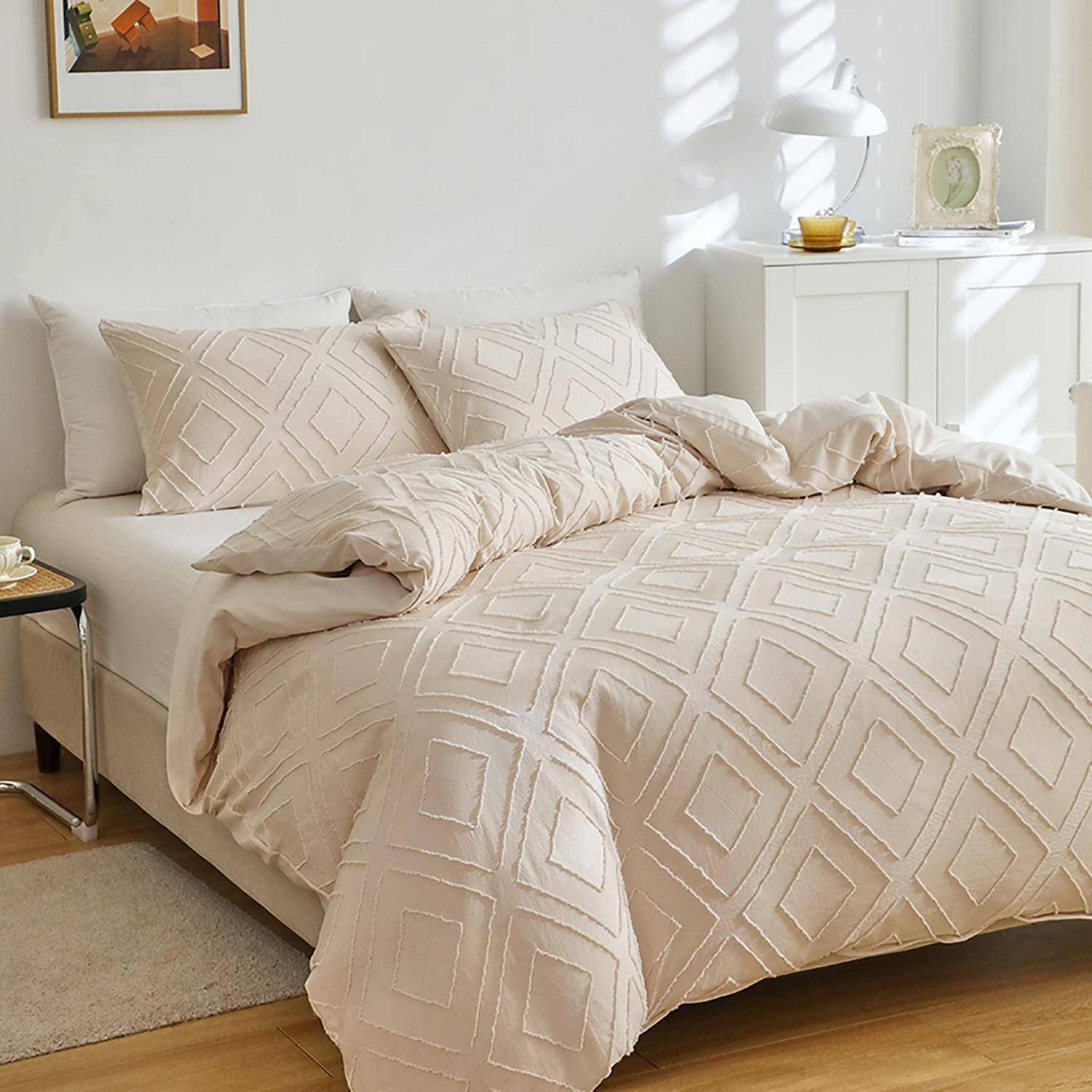 Sorgfältig gemachtes Bett mit beigefarbener geometrischer Bettwäsche in einem hellen Schlafzimmer, neben einem weißen Schrank und Wandkunst.