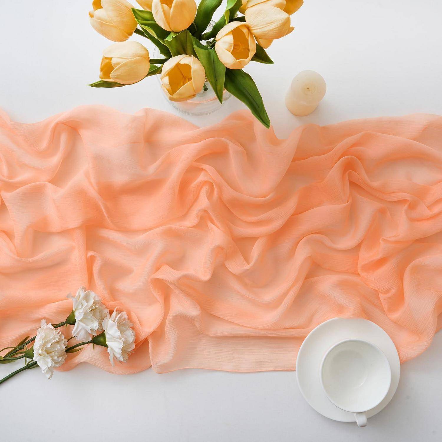 Pfirsichfarbener, durchsichtiger Stoff auf einer weißen Fläche mit gelben Tulpen, weißen Blüten und einer weißen Teetasse mit Untertasse als weiches, stilisiertes Flat-Lay.