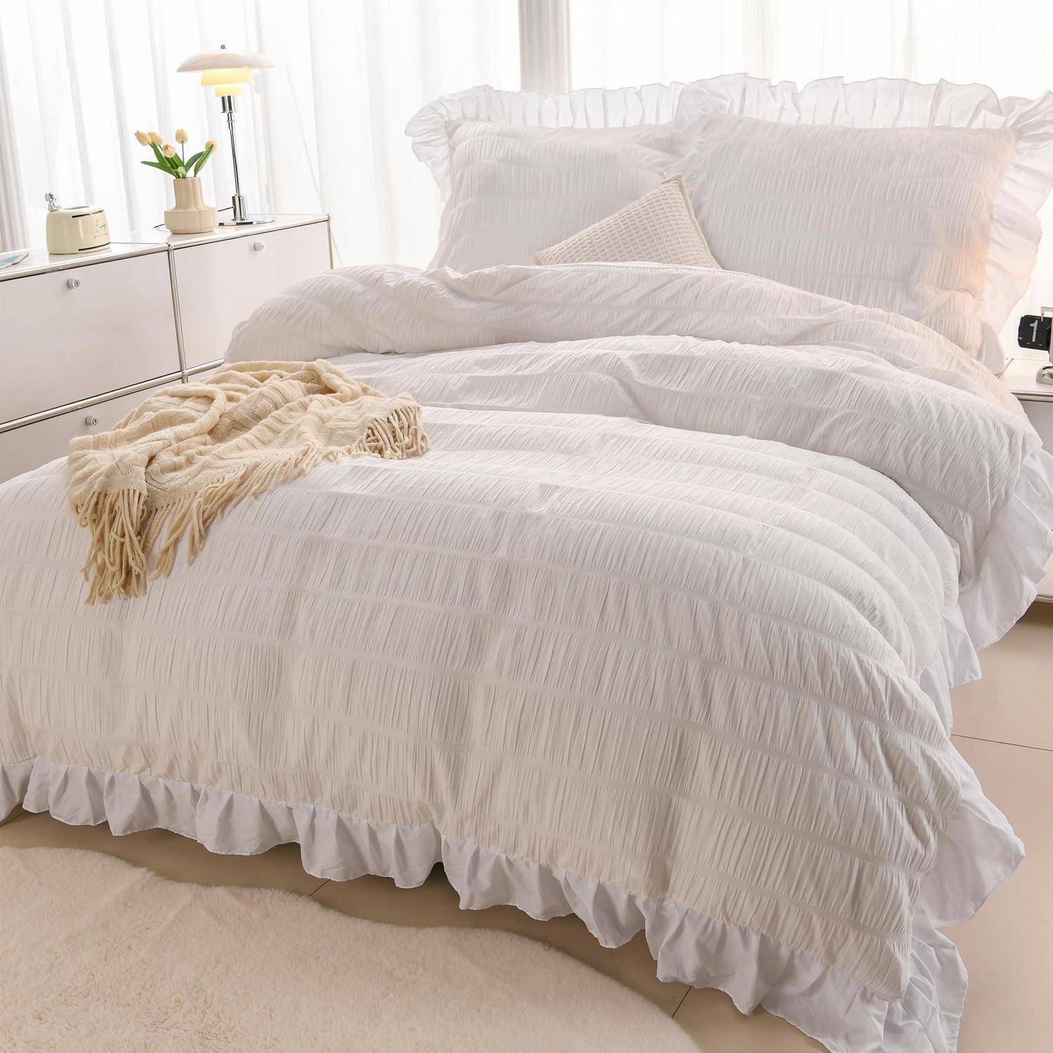 Weiße gerüschte Bettwäsche mit Kissen und eine cremefarbene Wurfdecke auf einem ordentlich gemachten Bett in einem hellen Schlafzimmer.