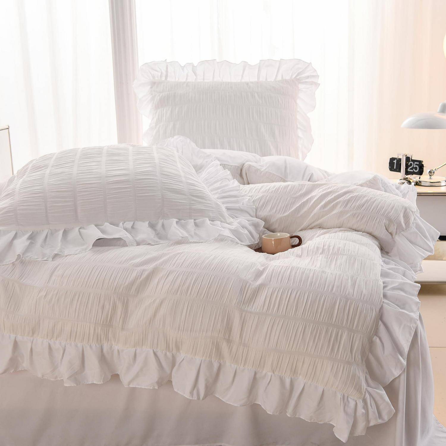 Weiße gekräuselte Bettwäsche auf einem unmachten Bett mit Kissen und einer Tasse, in einem hellen Schlafzimmer neben einem Nachttisch und einer Lampe.