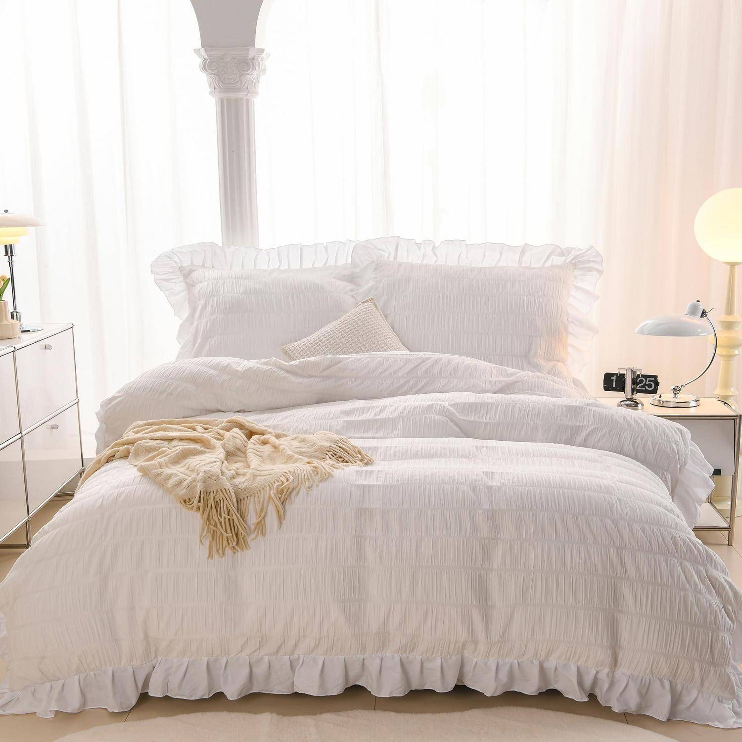Helles, weißes Schlafzimmer mit einem ungemachten Bett, das mit gekräuselter Bettwäsche, einer beigen Decke und Nachttischlampen neben großen transparenten Vorhängen versehen ist.