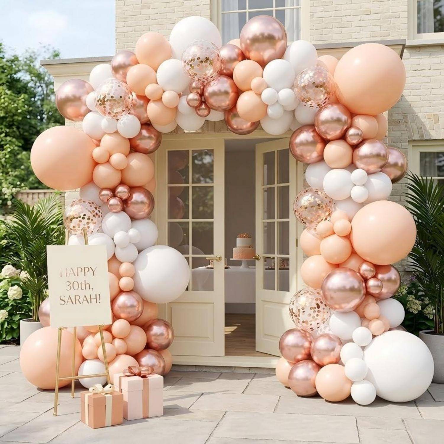 Pfirsich-, weiß- und rosegoldfarbene Ballongirlande um einen Türrahmen mit einem Schild, auf dem "Happy 30th, Sarah!"" steht, und Geschenkboxen für eine Geburtstagsfeier.