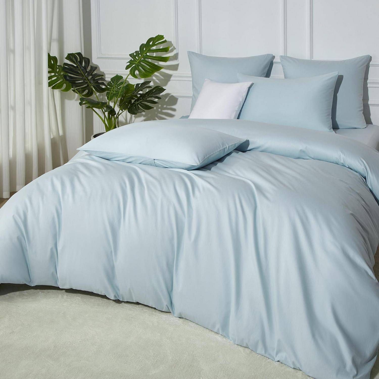 Hellblaues Bett mit passenden Kissen und Bettdecke in einem hellen, minimalistischen Schlafzimmer neben einer großen grünen Pflanze.