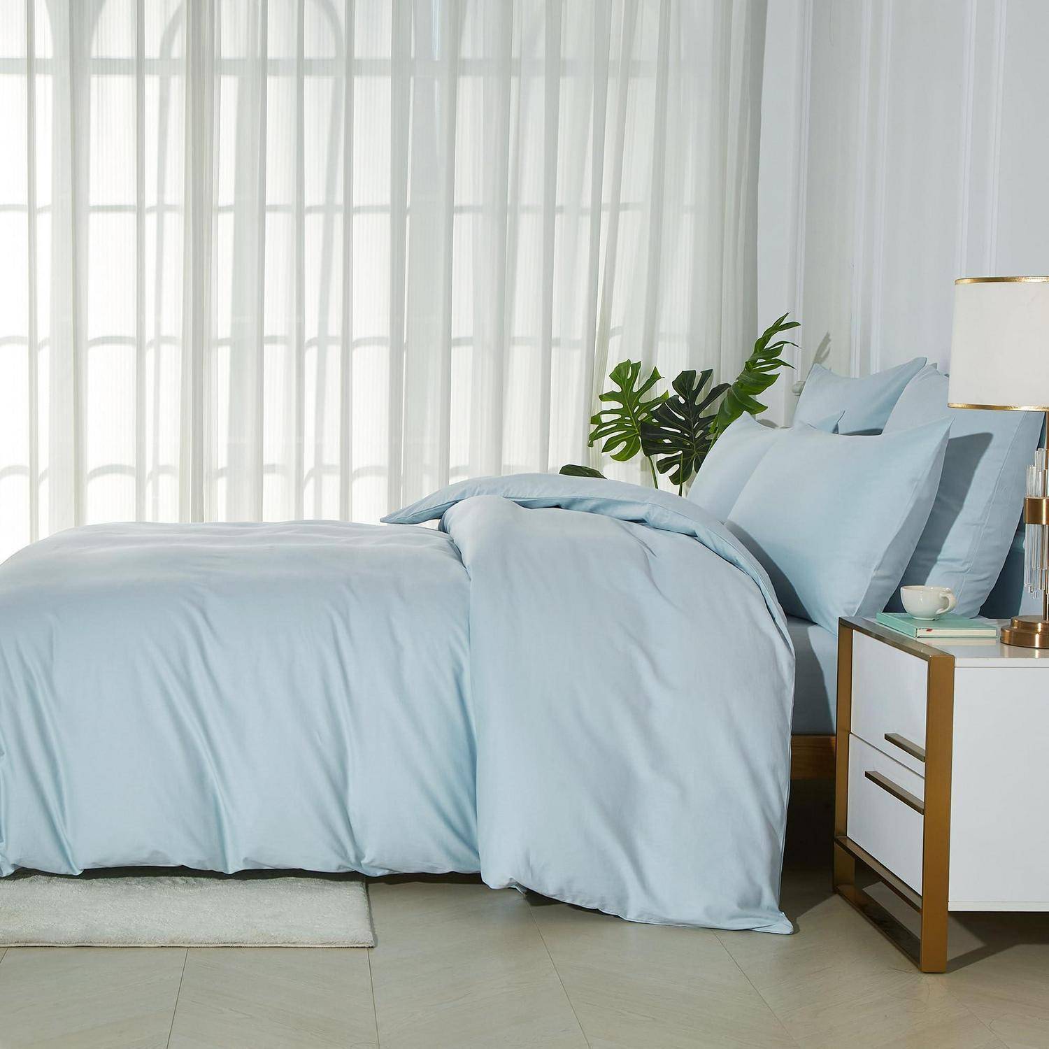 Ein ordentlich gemachtes Bett mit hellblauer Bettwäsche neben einem weißen Nachttisch und einer Lampe in einem hellen, modernen Schlafzimmer.