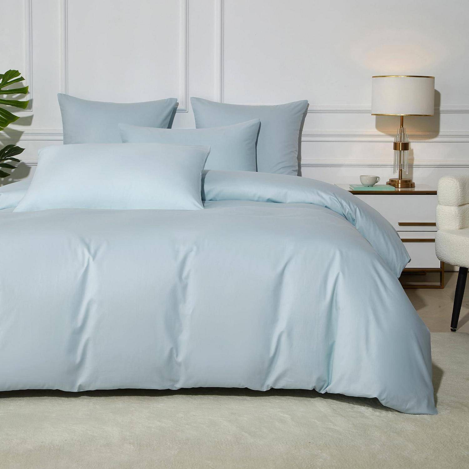 Ein ordentlich gemachtes Bett mit hellblauer Bettwäsche und Kissen in einem minimalistischen weißen Schlafzimmer, neben einem Nachttisch mit Lampe und Tasse.