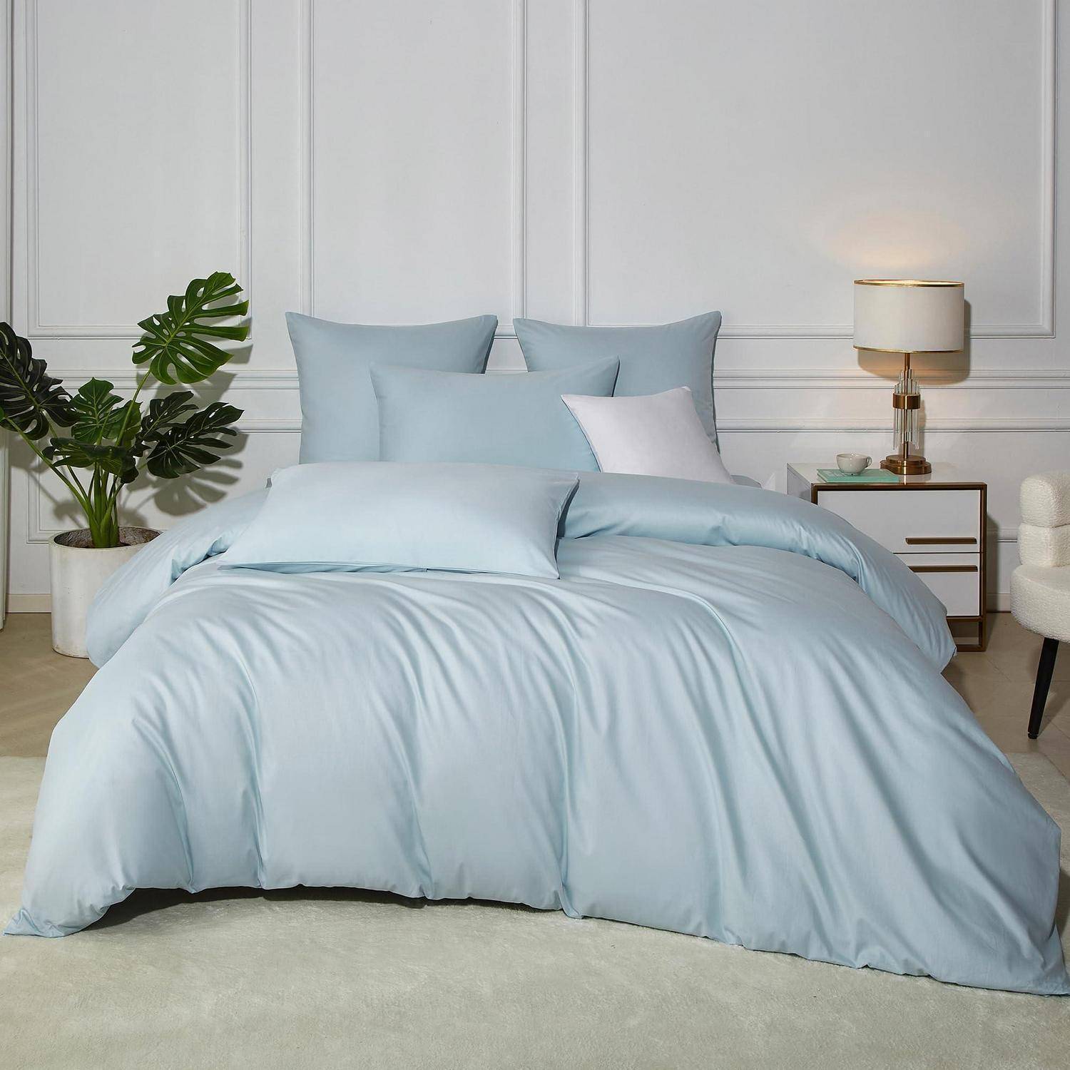 Hellblaue Bettwäsche mit Kissen auf einem ordentlich gemachten Bett in einem minimalistischen Schlafzimmer, mit einer Lampe, einer Pflanze und einem kleinen Nachttisch.