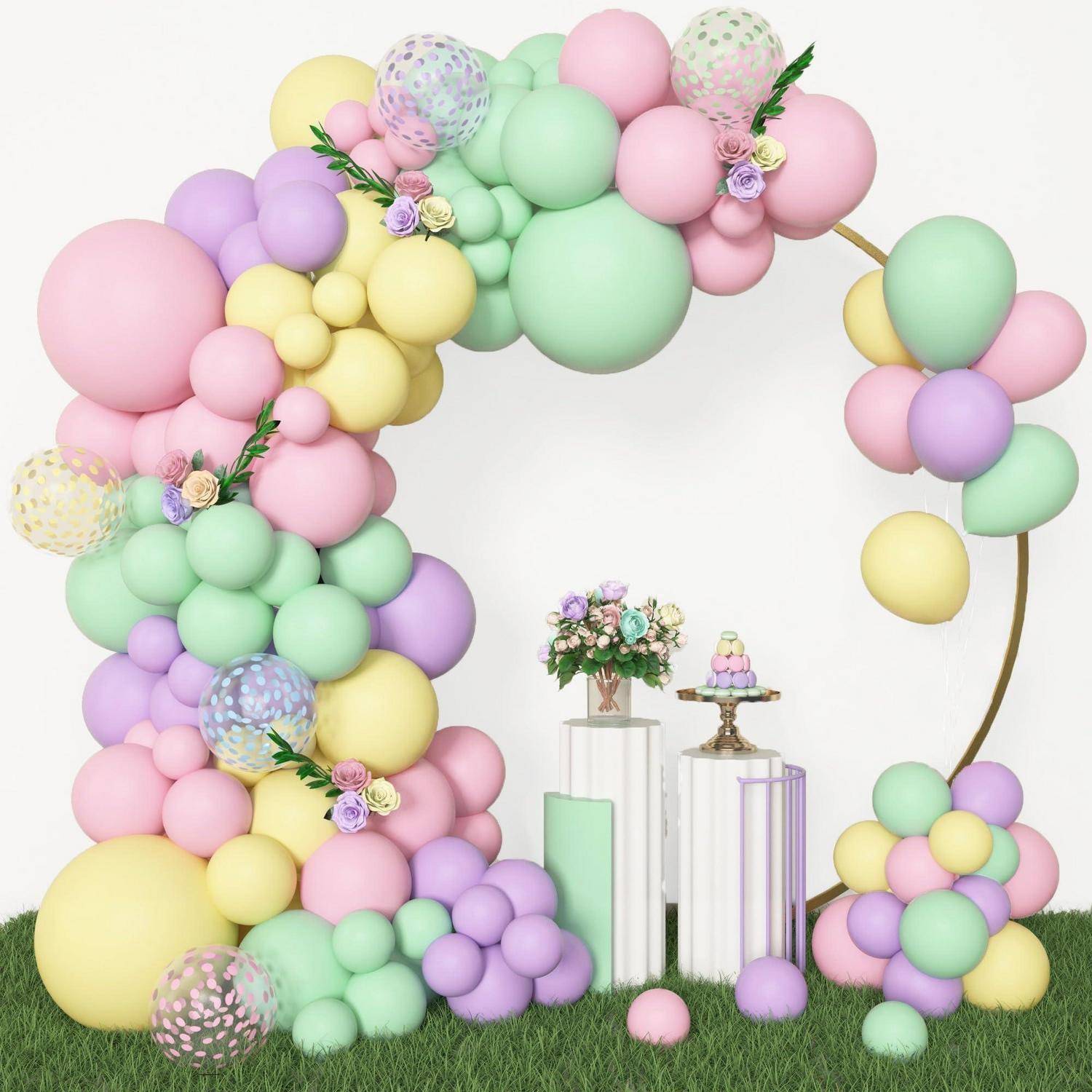 Pastellfarbener Ballonbogen und Ballontrauben als Rahmung einer kleinen Kuchenausstellung auf weißen Sockeln, aufgestellt auf grünem Rasen als festlicher Partyhintergrund.