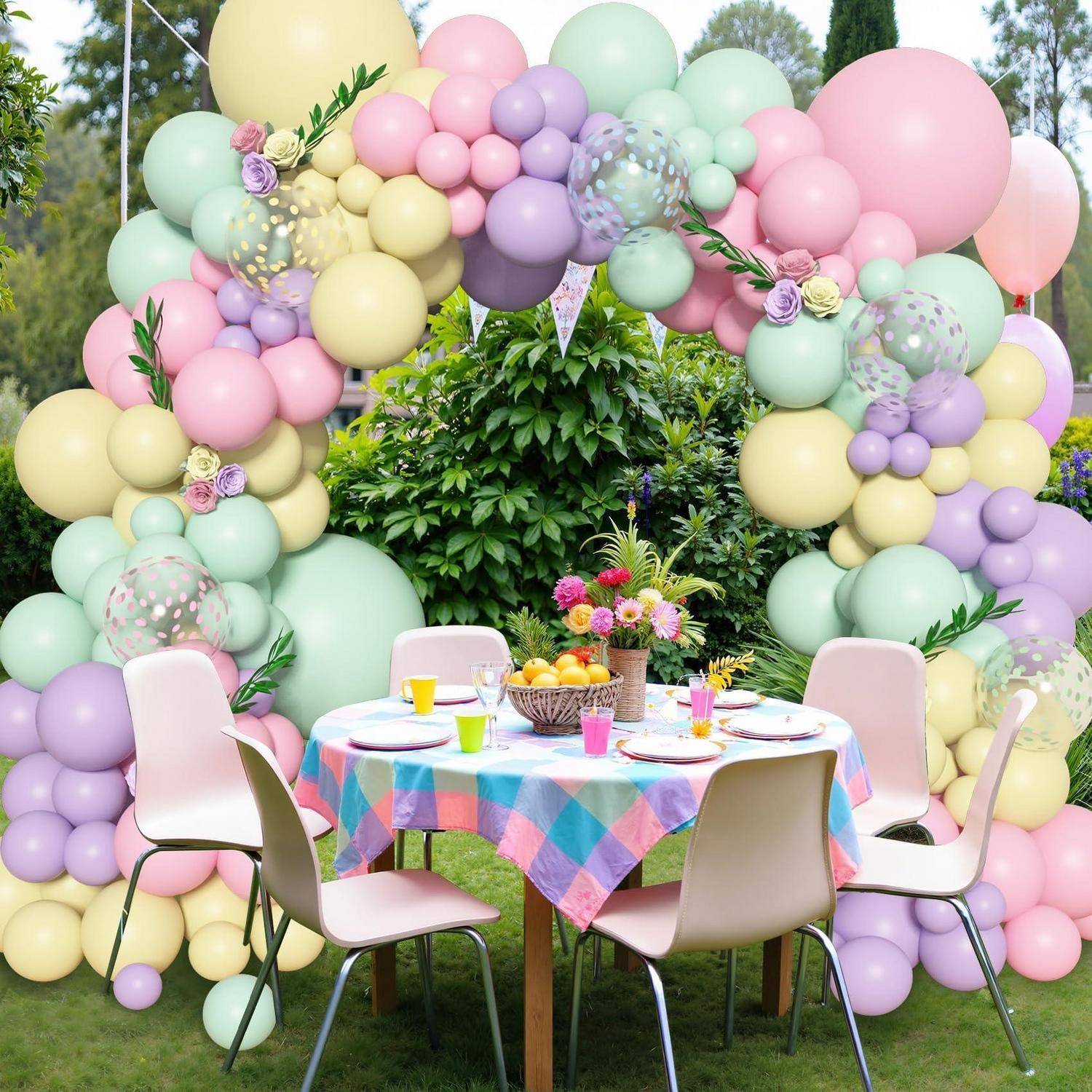 Gartenparty-Tisch für vier Personen unter einem pastellfarbenen Ballonbogen, mit Blumen, Obst und bunten Platzdeckchen auf einem Rasen.