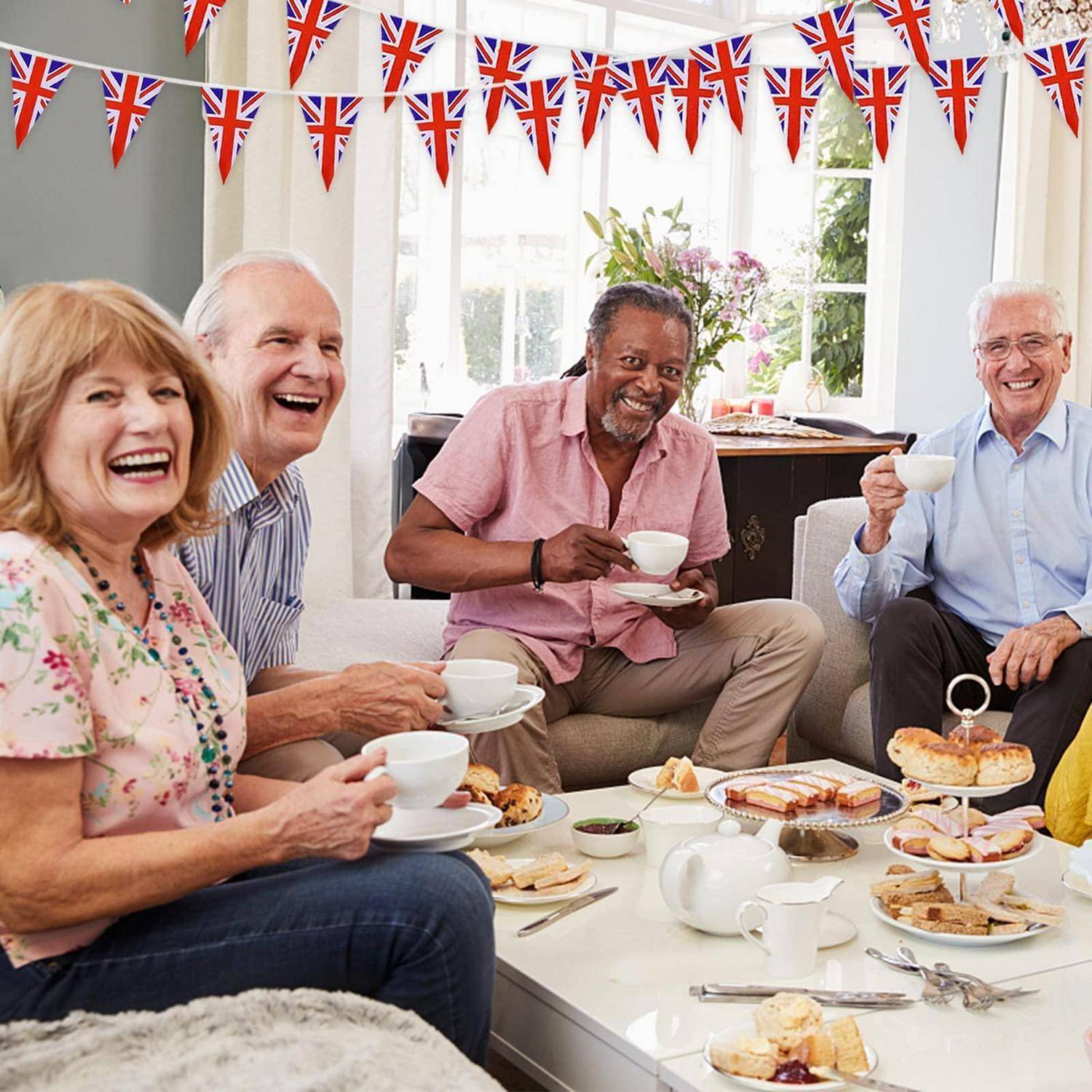 Lächelnde ältere Erwachsene teilen Tee und Kuchen bei einer fröhlichen Innenveranstaltung, die mit Union-Jack-Girlanden geschmückt ist.
