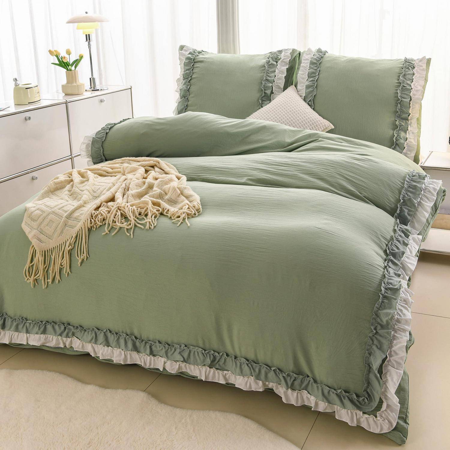 Ordentlich gemachtes Bett mit graugrüner gerüschter Bettwäsche und Kissen, eine cremefarbene Wurfdecke, neben einer weißen Kommode in einem hellen Schlafzimmer.