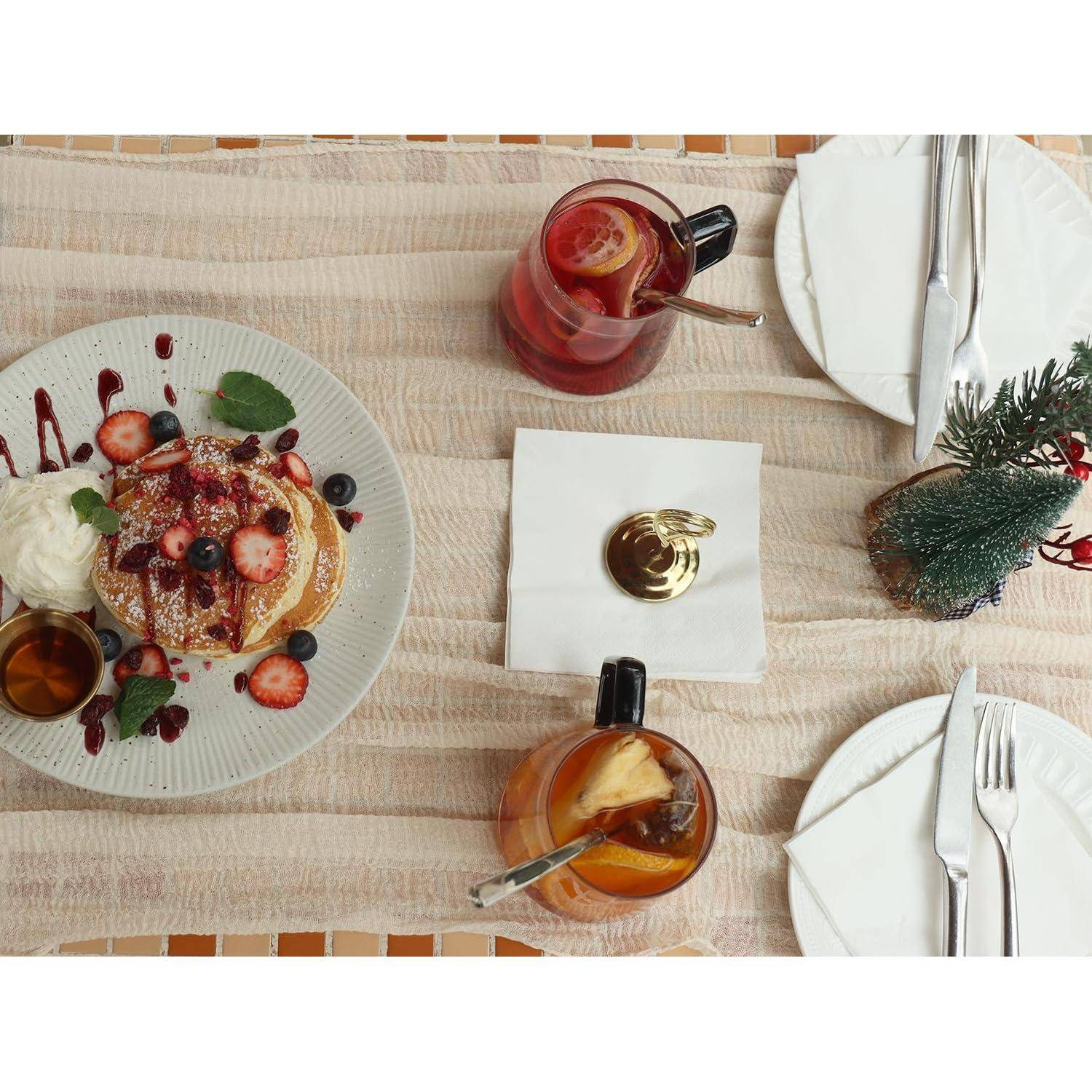 Draufsicht auf einen Brunchtisch mit Pfannkuchen, die mit Erdbeeren, Schlagsahne und Sirup belegt sind, daneben zwei Getränke, Teller, Besteck und ein kleiner dekorativer Baum.