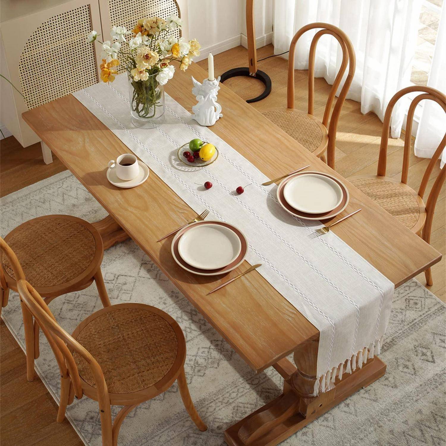 Tisch im hellen Zimmer decken, mit Holzstühlen, weißem Läufer, Tellern, Tassen, Obst und Blumen.